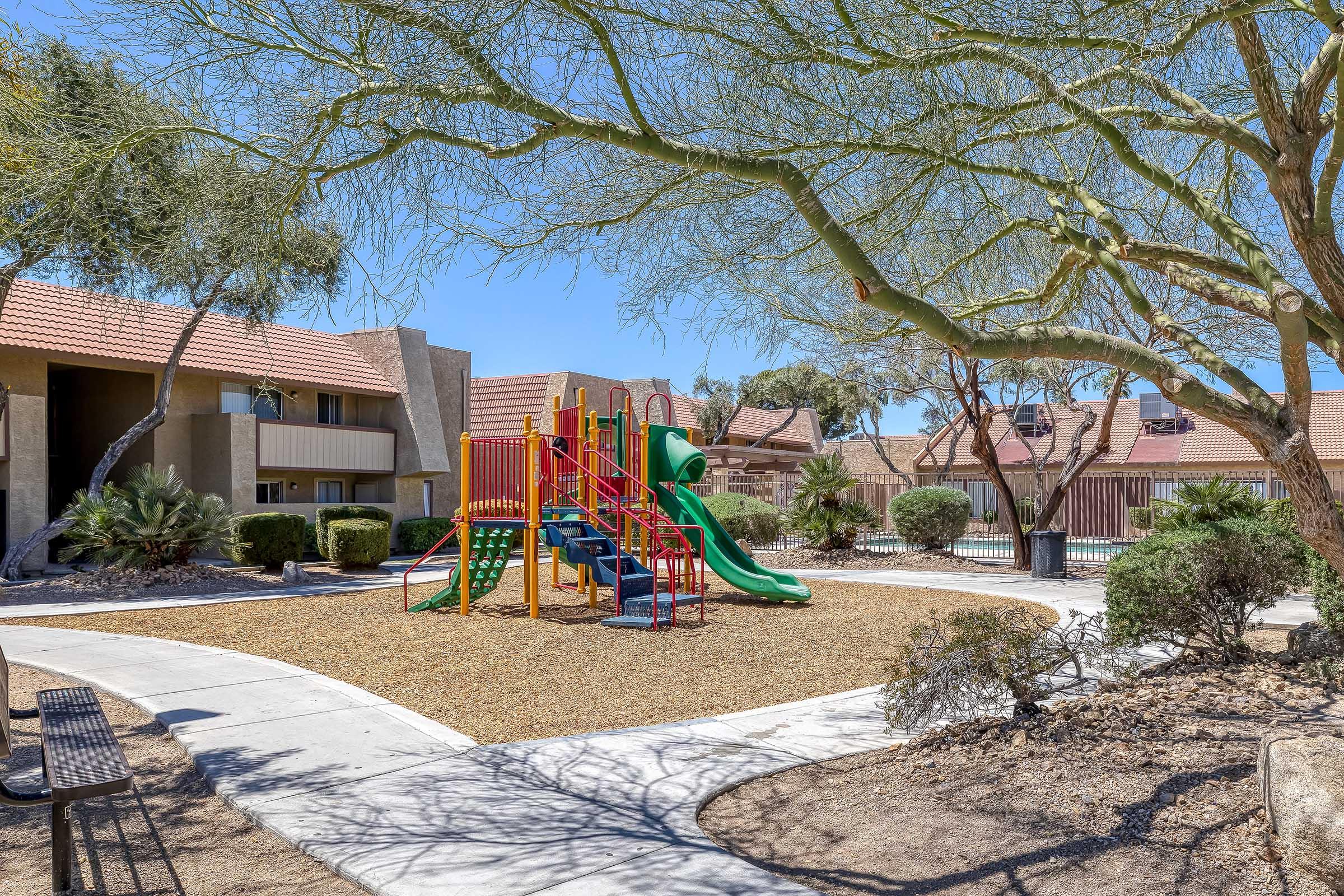 A playground featuring colorful climbing structures and slides, surrounded by a gravel area and desert vegetation. In the background, there are residential buildings with red tiled roofs. The scene is set under a clear blue sky with some trees providing shade.
