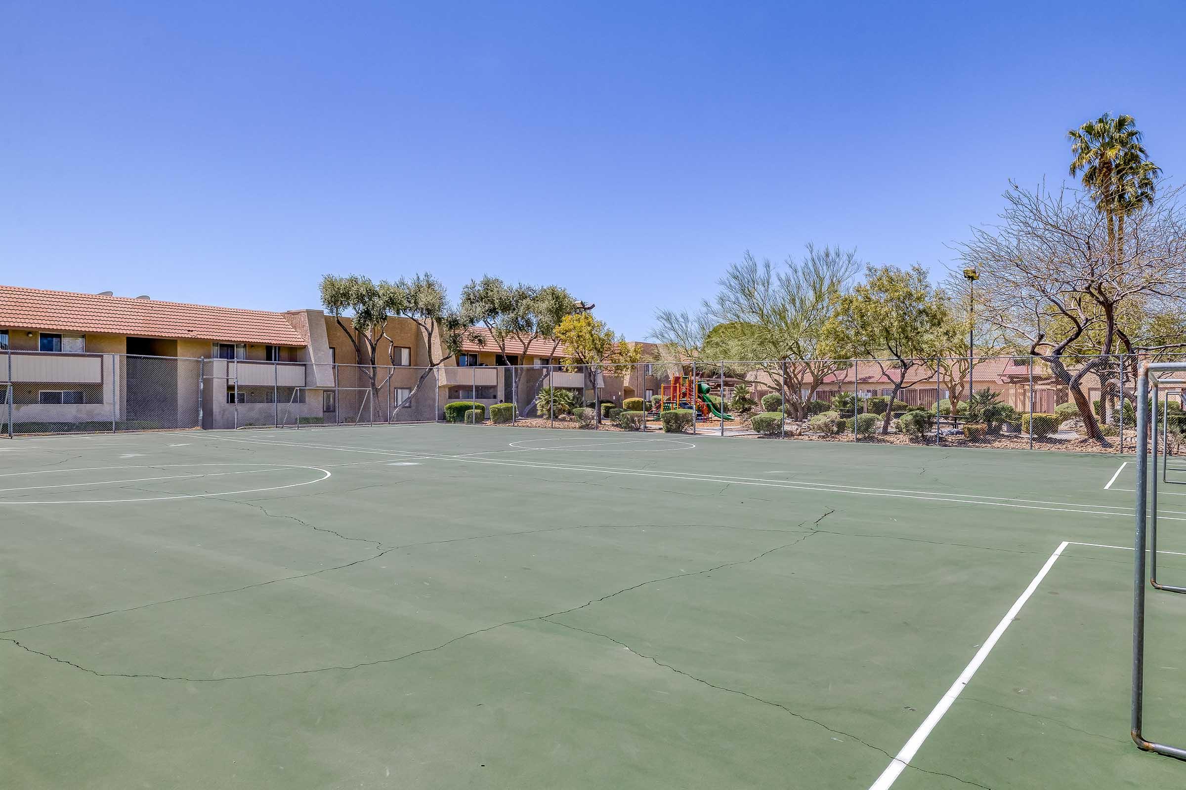 A vacant outdoor basketball court surrounded by desert landscaping and residential buildings. The court has visible lines, a few cracks, and no players present. In the background, there are trees, shrubs, and a playground area. Clear blue sky overhead with no clouds.