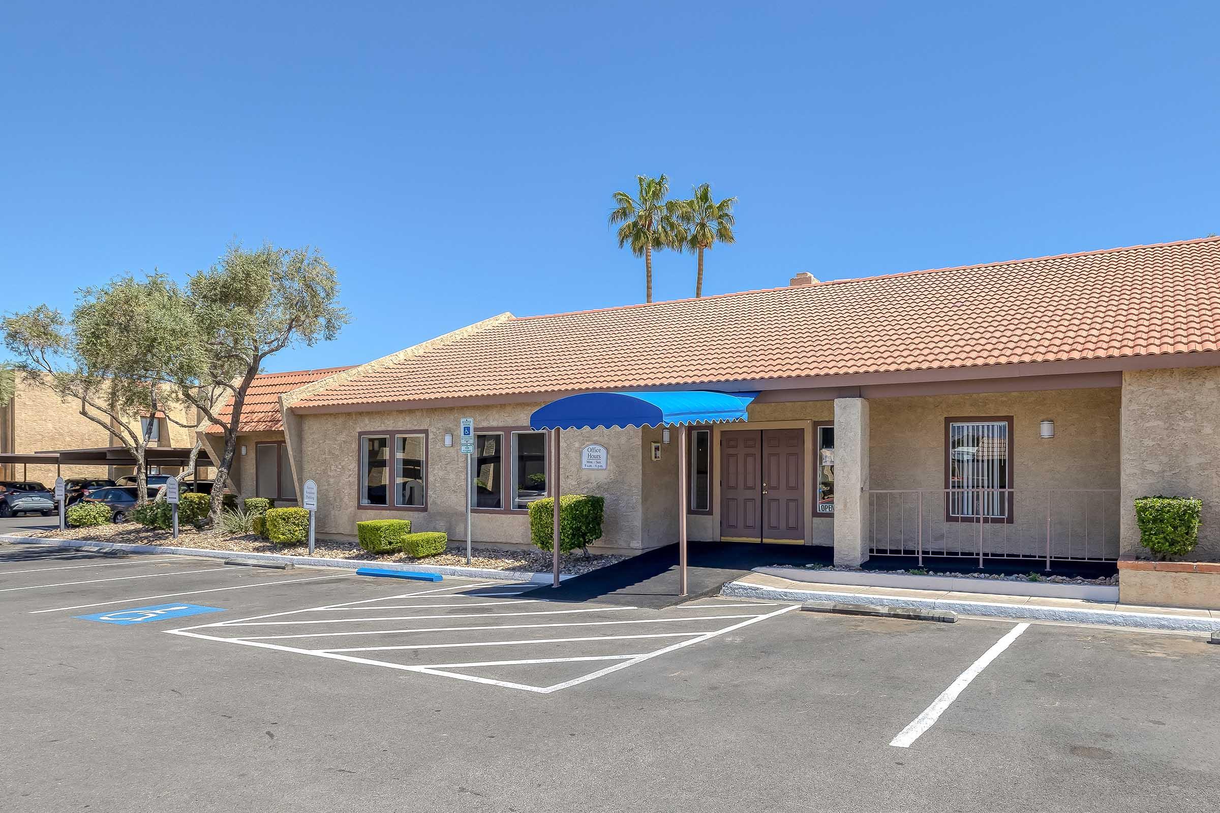 A single-story building with a reddish-brown tiled roof and a blue awning. There are two doors at the front, flanked by small shrubs. The parking lot in front is marked with painted lines. Palm trees can be seen nearby, set against a clear blue sky.