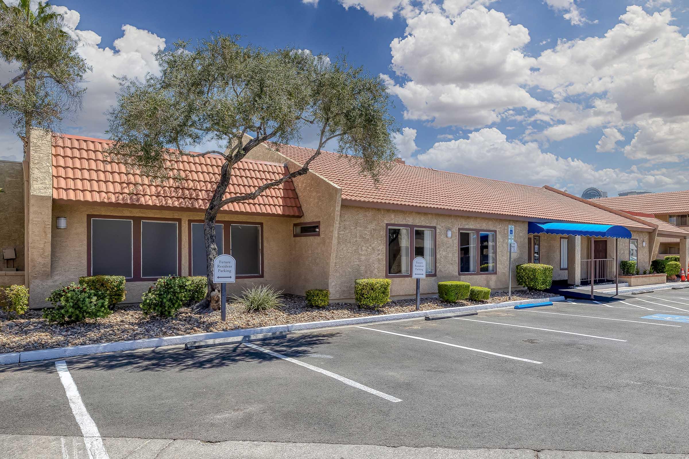 A single-story building with a tiled roof, surrounded by a landscaped area featuring shrubs and small trees. There are several parking spaces in front, and a blue awning covers the entrance. The sky is partly cloudy, creating a bright and inviting atmosphere.
