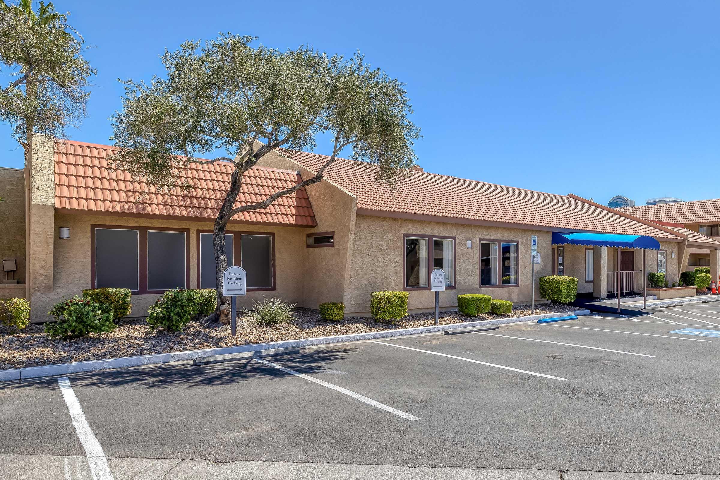 A single-story building with a red tile roof and beige stucco exterior. There are several large windows, a blue awning at the entrance, and neatly trimmed bushes in the front. The parking lot is marked with spaces, and the area is sunny with a clear blue sky.