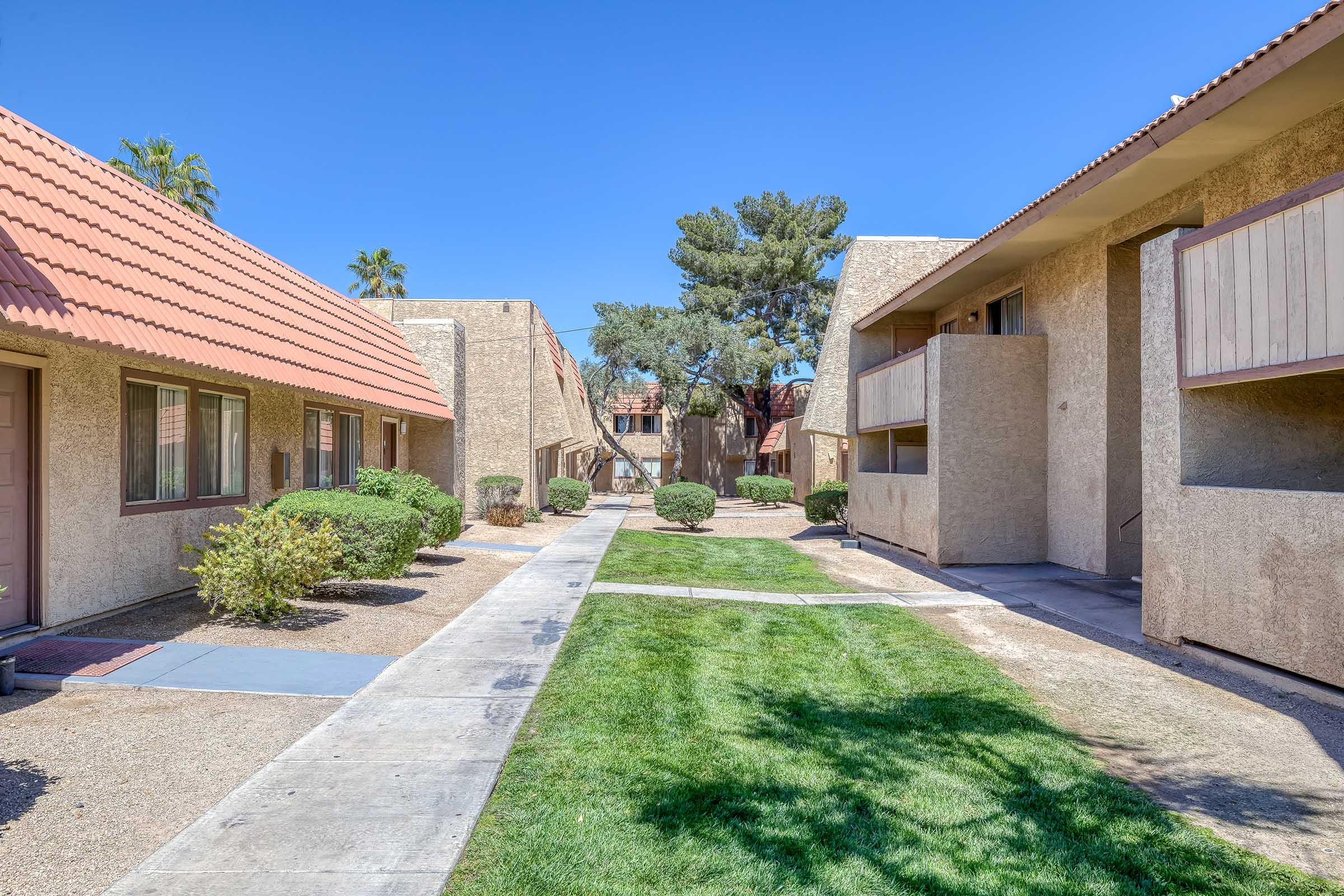 A pathway lined with well-maintained grass and small shrubs connects two sets of apartment buildings. The buildings feature a textured tan exterior and terracotta roofs, set against a clear blue sky. Palm trees are visible, adding to the pleasant outdoor atmosphere.