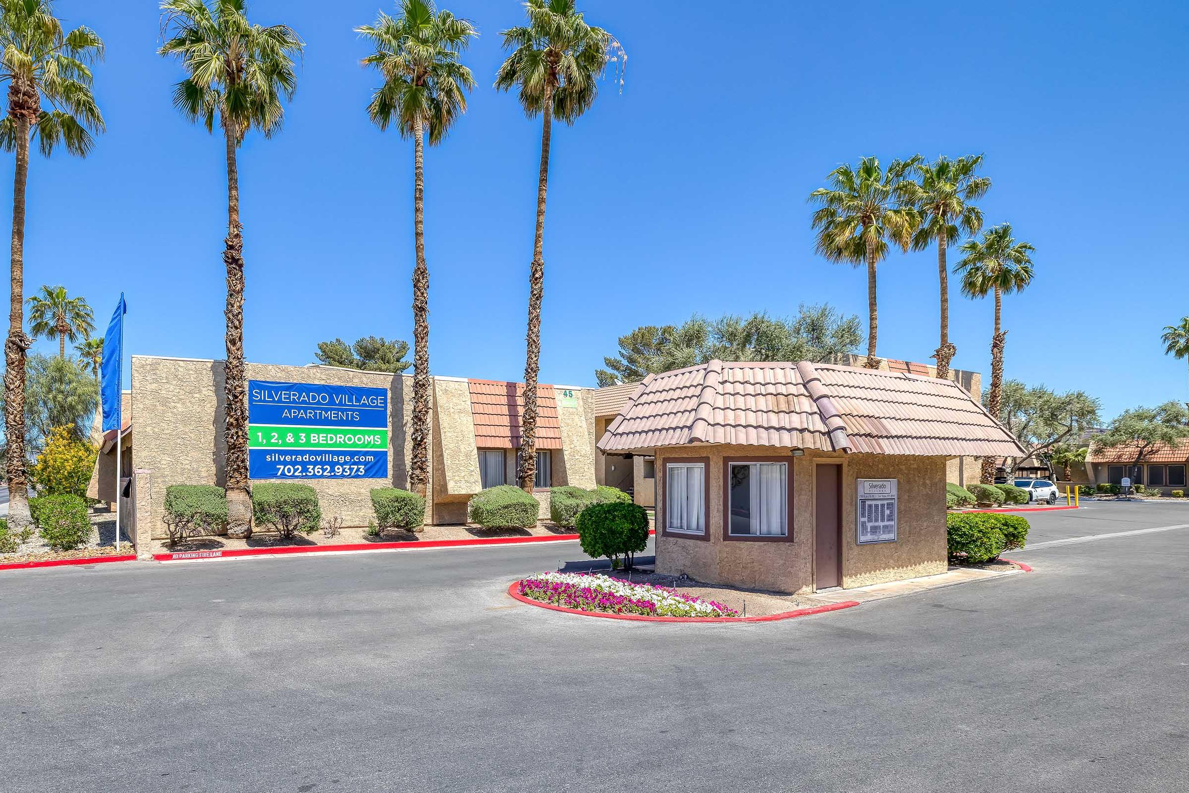 Entrance to Silverado Village Apartments with palm trees lining the pathway. The building features a sign indicating 1, 2, and 3-bedroom apartments available. Colorful flower beds enhance the landscaping, and the clear blue sky completes the welcoming atmosphere.