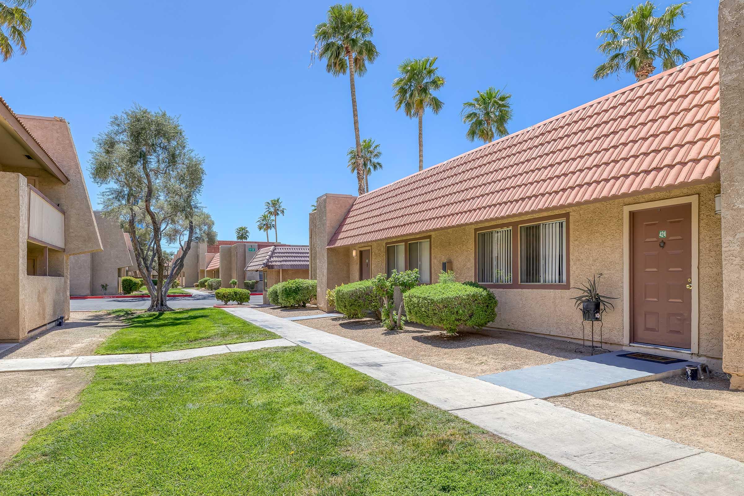 A sunny view of a residential apartment complex featuring beige buildings with red tiled roofs, palm trees, and manicured shrubs. A concrete pathway runs through the lawn, leading to the entrance of one of the units. The scene conveys a warm, inviting atmosphere typical of a Southwest setting.