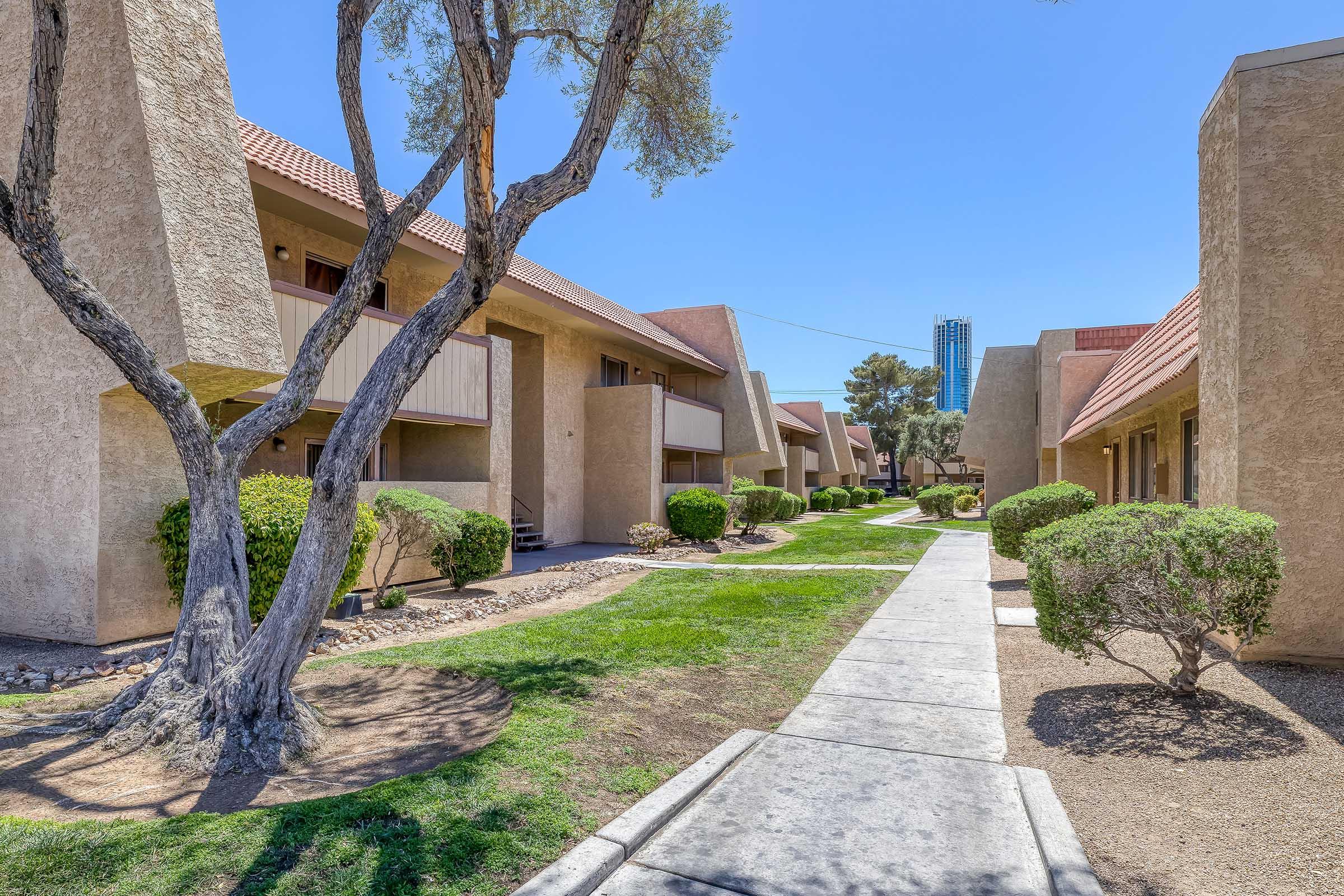 A view of an apartment complex featuring two-story buildings with beige exteriors and reddish rooftops. Lush green lawns and neatly trimmed shrubs line the walkway, which leads to the entrance of the apartments. A few trees provide shade, and the sky is clear and blue.