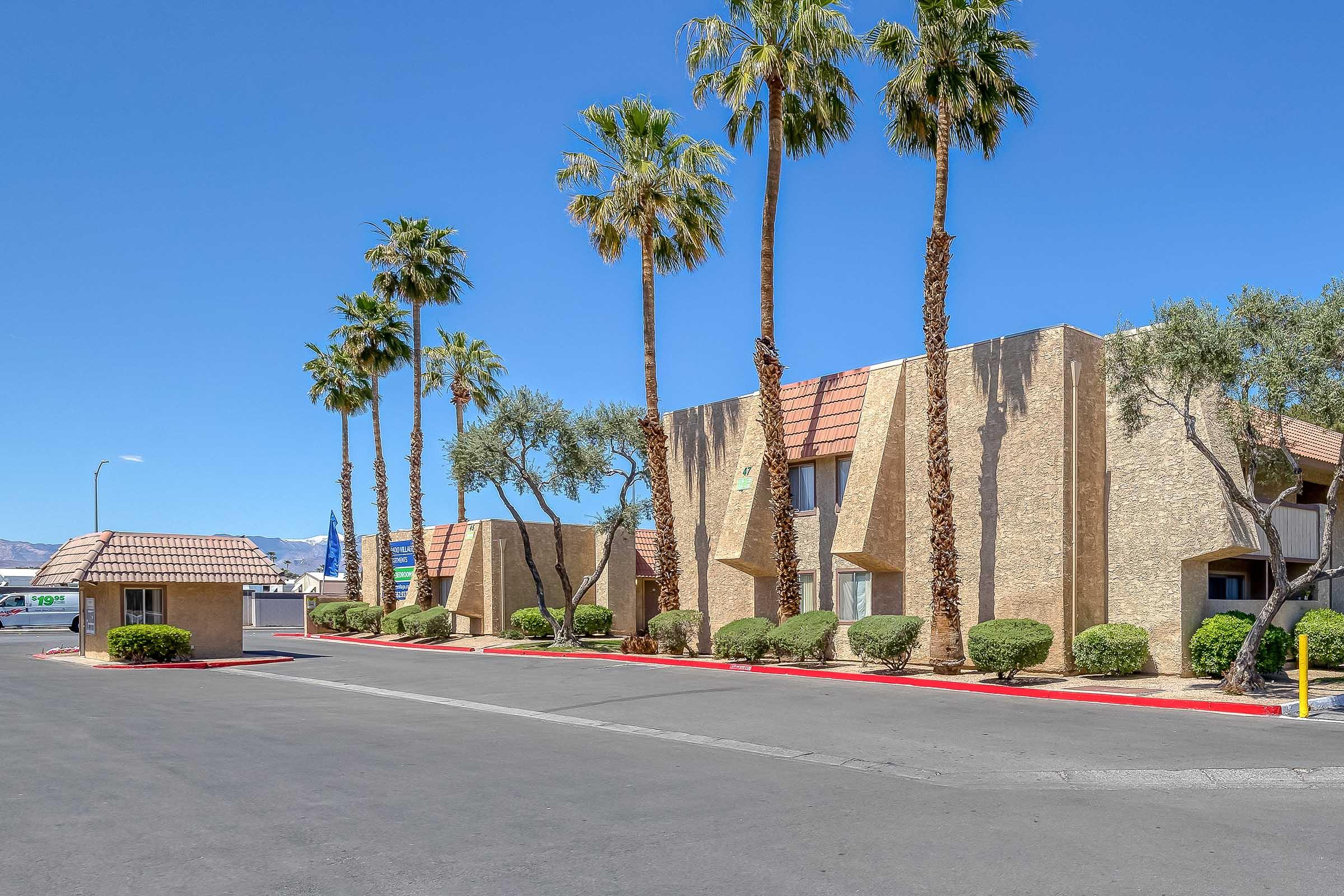 A view of a landscaped area featuring several tall palm trees and contemporary buildings with beige exteriors. The setting is bright and sunny, with a clear blue sky and a smooth asphalt road leading to the entrance of the property.