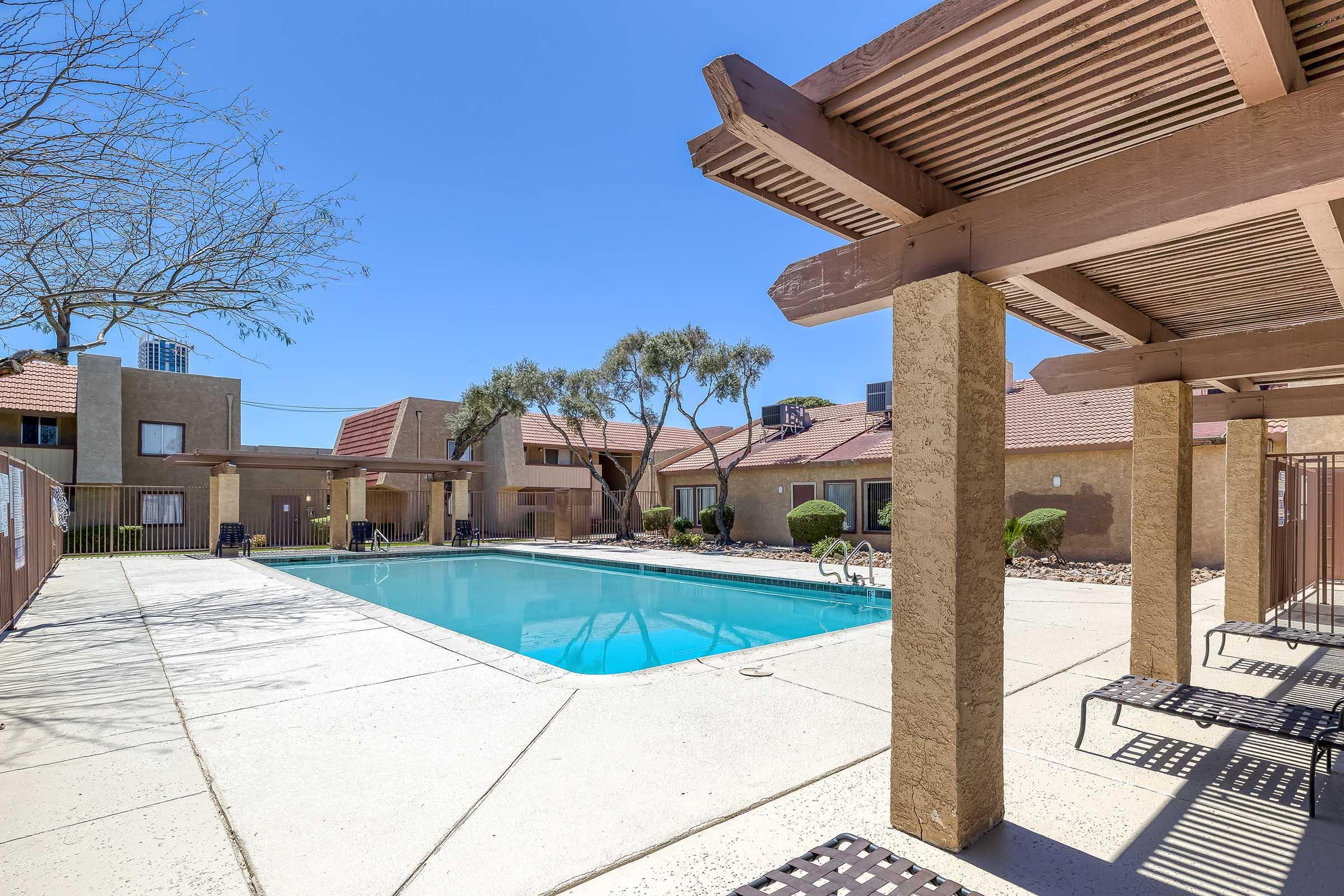 A bright outdoor pool area in a residential complex, surrounded by well-maintained landscaping and cozy seating under pergolas. The clear blue sky complements the inviting pool, while buildings with terracotta roofs are visible in the background.