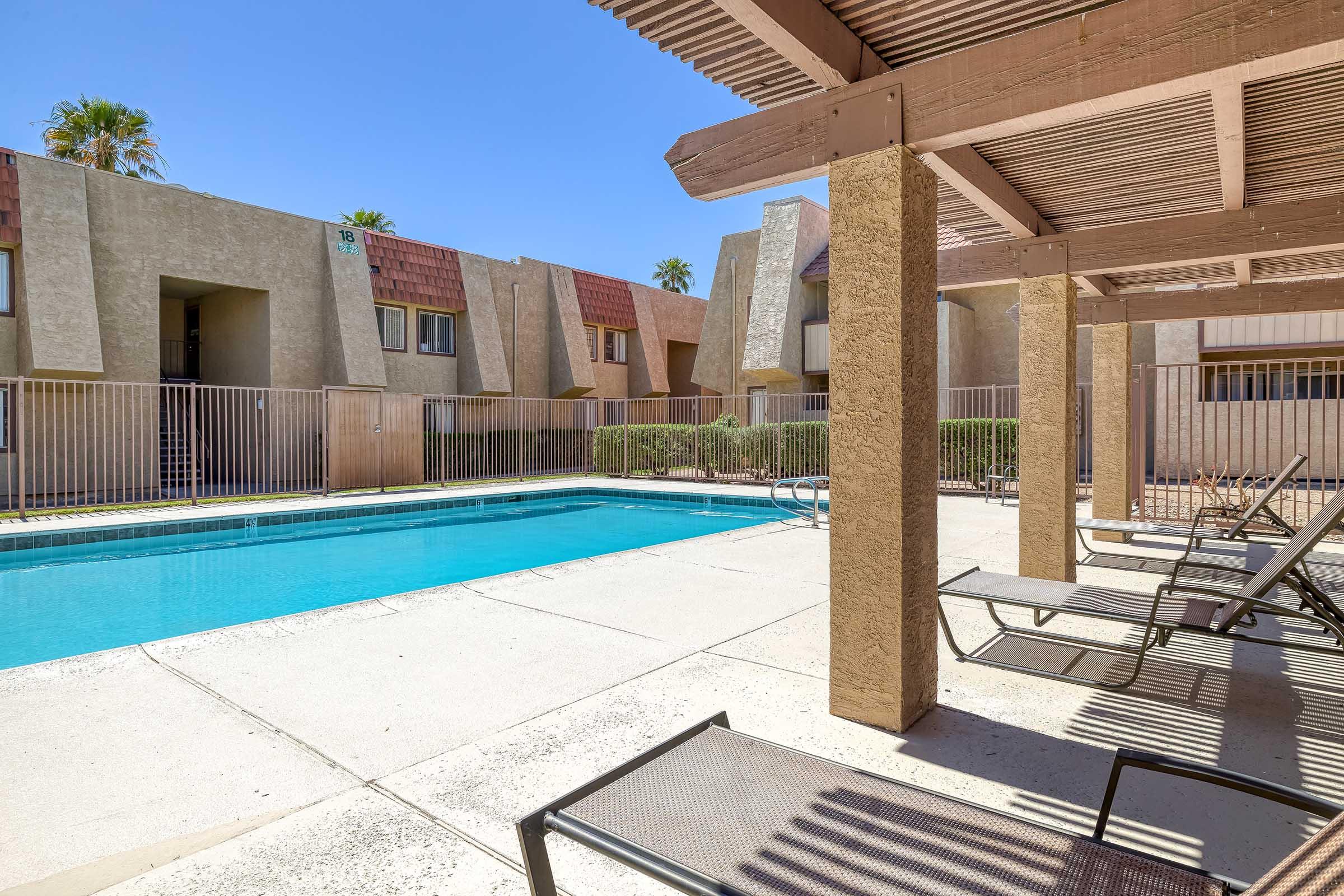 A clear blue swimming pool surrounded by lounge chairs, set in a courtyard of beige apartment buildings. The area is sunny with a blue sky and palm trees in the background. A fence encloses the pool area, providing a private and relaxing atmosphere.