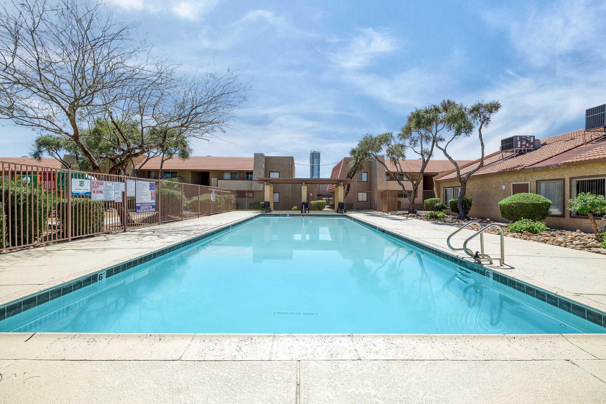 A clear swimming pool surrounded by a gated area, with palm trees and shrubs in the background. The pool area features lounge chairs and is bordered by two apartment buildings. Bright blue sky and a few clouds above, giving a sunny, inviting feel to the scene.
