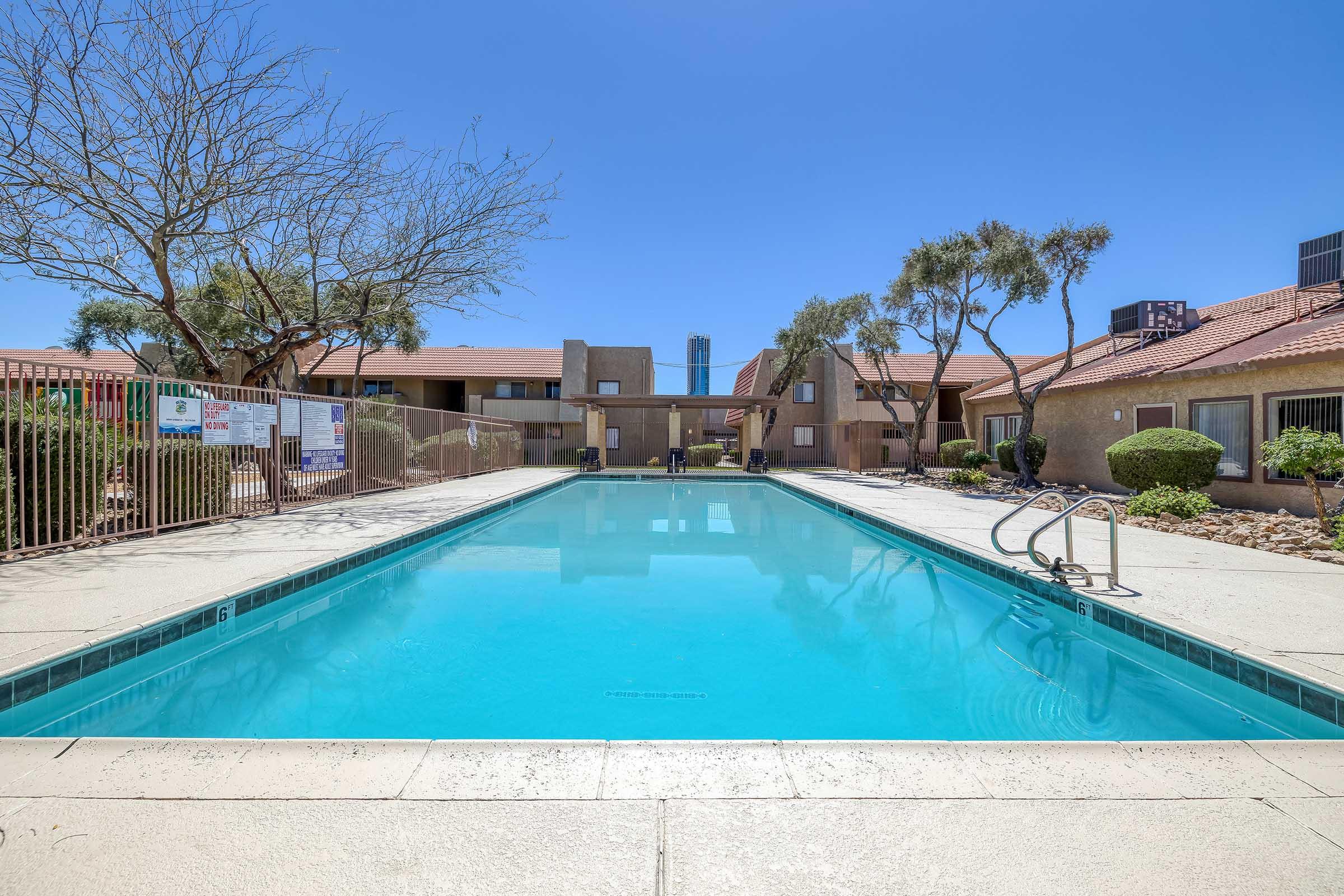 A clear blue swimming pool surrounded by a fence, with lounge chairs on one side. In the background, there are residential buildings with a red-tiled roof and a few trees. The sky is bright blue, indicating a sunny day.