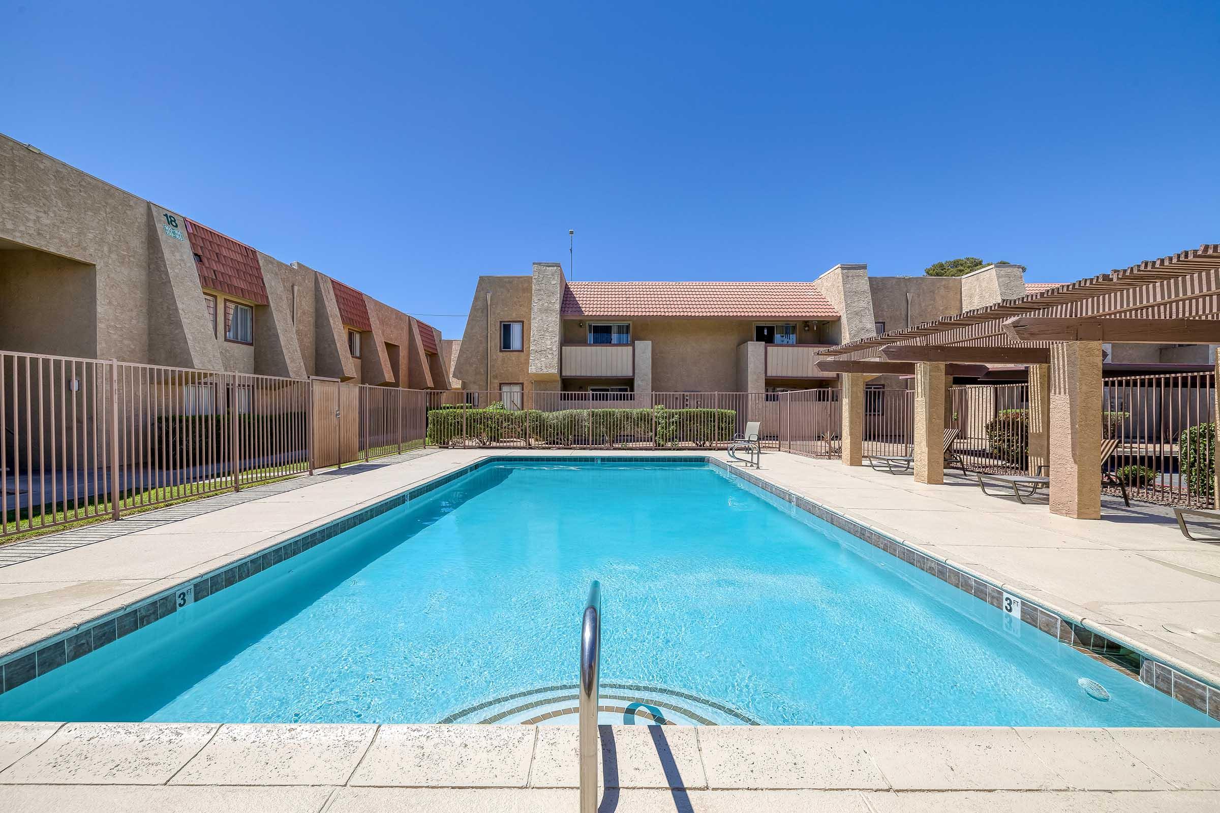 A clear blue swimming pool surrounded by beige apartment buildings and green landscaping. The pool area features a fenced perimeter and shaded seating areas, with a bright blue sky overhead.