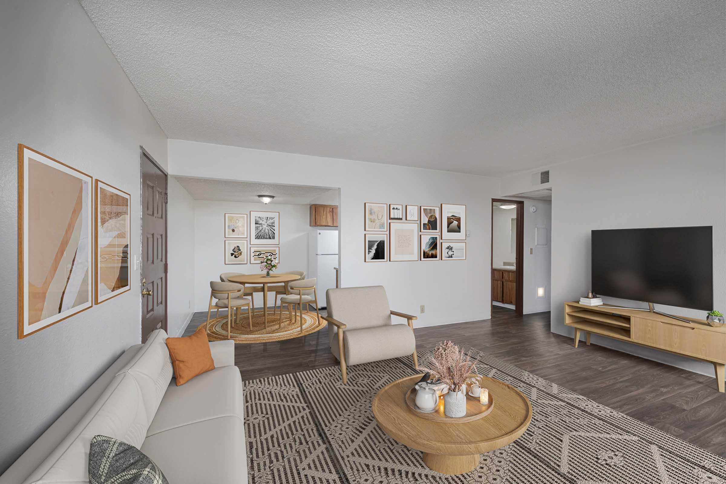 A modern living room featuring a light-colored sofa with an orange pillow, a round wooden coffee table with decorative items, and a large flat-screen TV on a wooden console. The walls are adorned with framed artwork, and a dining area with a round table and chairs is visible in the background. The space has a neutral color palette and textured flooring.