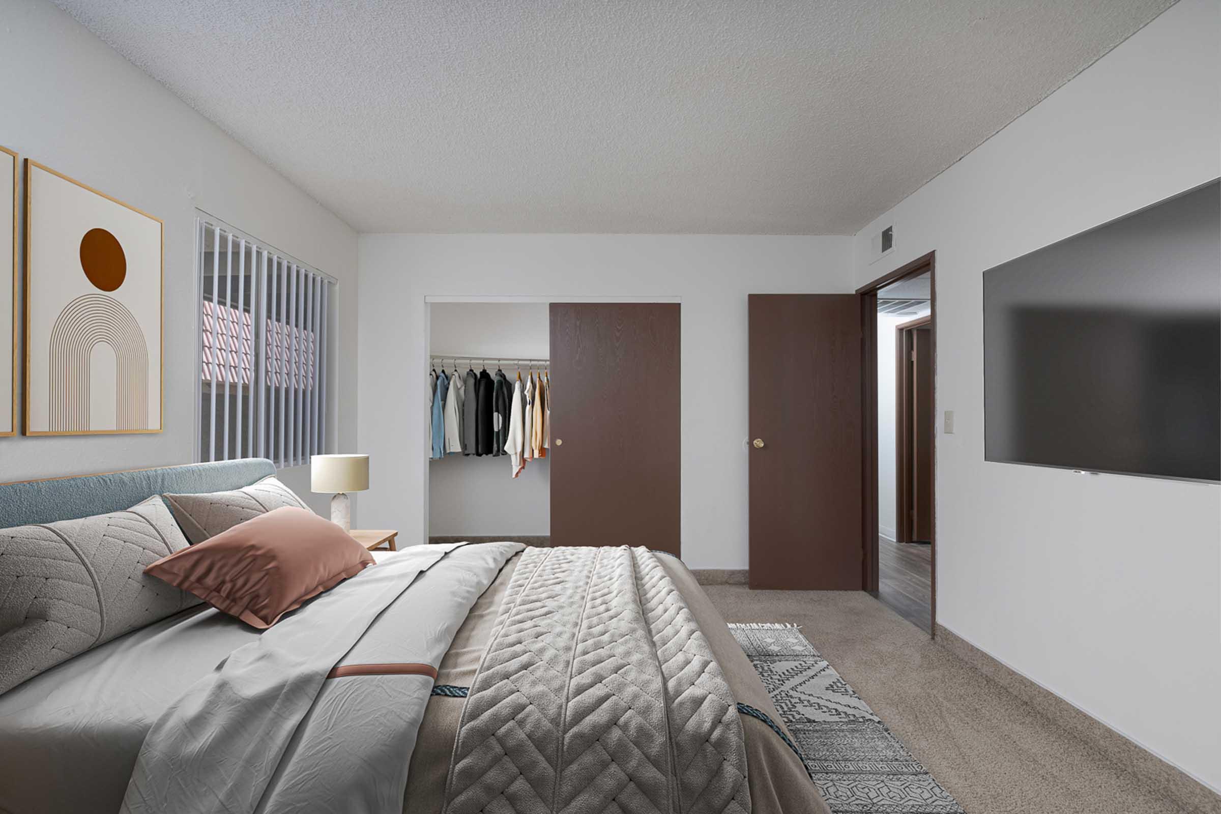 A modern bedroom featuring a neatly made bed with gray linens and decorative pillows. A lamp sits on a bedside table. The room includes a closet with hanging clothes, a closed wooden door, and a wall-mounted TV opposite the bed. The walls are light-colored, and there’s a textured rug on the floor.