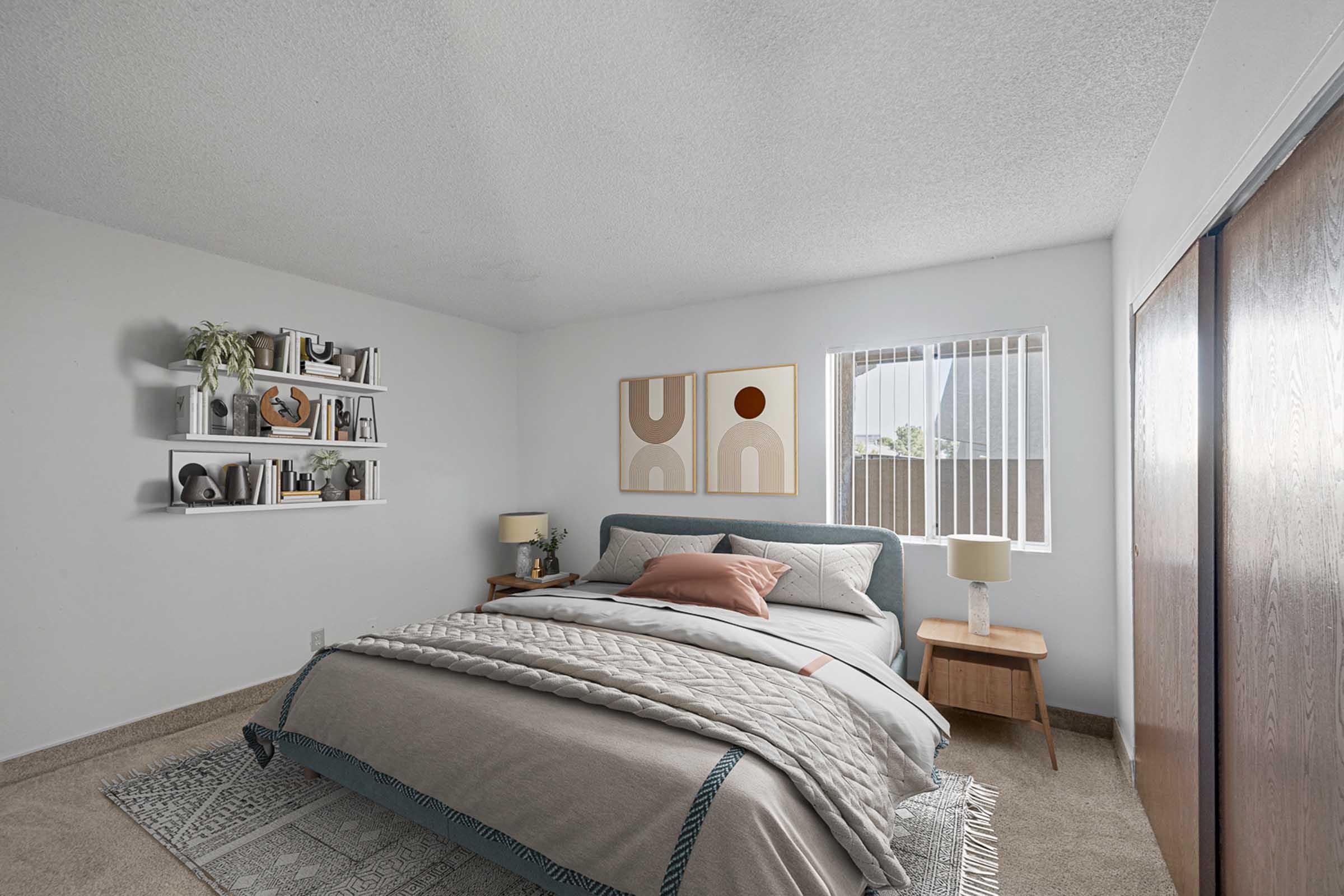 Cozy bedroom featuring a neatly made bed with a light gray blanket and decorative pillows, a small wooden nightstand with a lamp, and wall-mounted shelves displaying various decor items. Bright natural light enters through a window, illuminating the softly colored walls and carpeted floor.