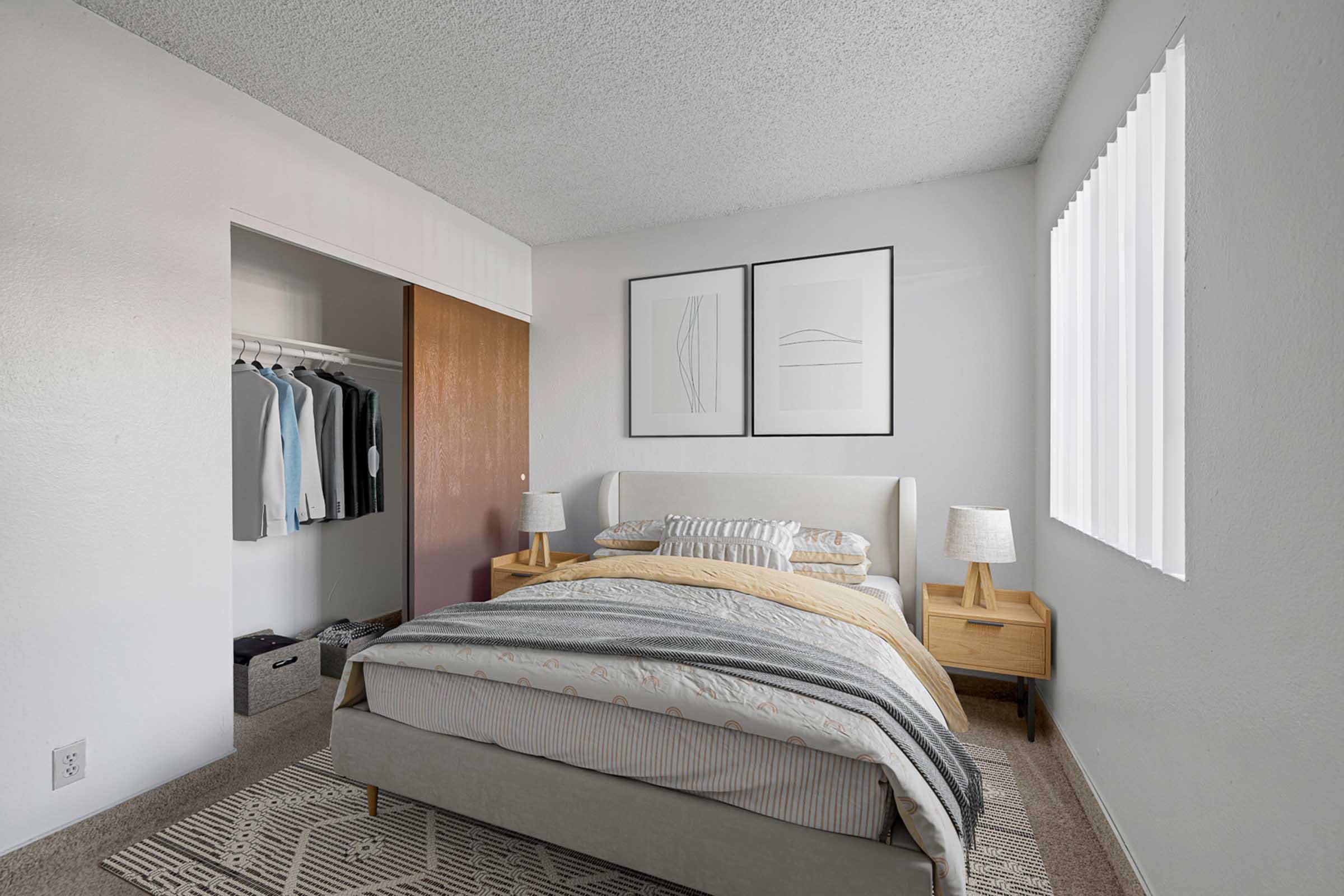 A cozy bedroom featuring a light-colored bed with decorative pillows and a yellow blanket. There are two wooden bedside tables with lamps, a closet with hanging clothes, and minimalistic artwork on the walls. Natural light is coming in through a window with vertical blinds.