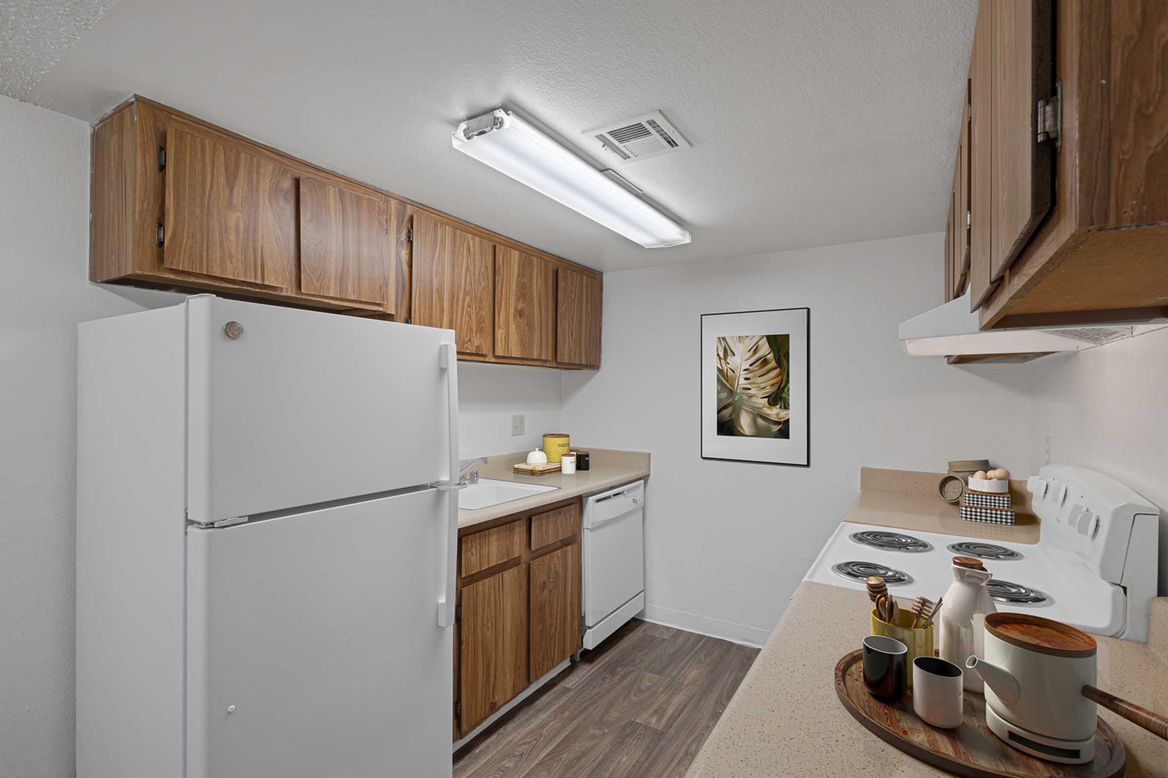 A modern kitchen featuring wooden cabinets, a white refrigerator, a sink with a dishwasher, and a white stove. The countertop is decorated with kitchenware and a framed botanical print hangs on the wall. The lighting is bright, enhancing the clean and organized look of the space.