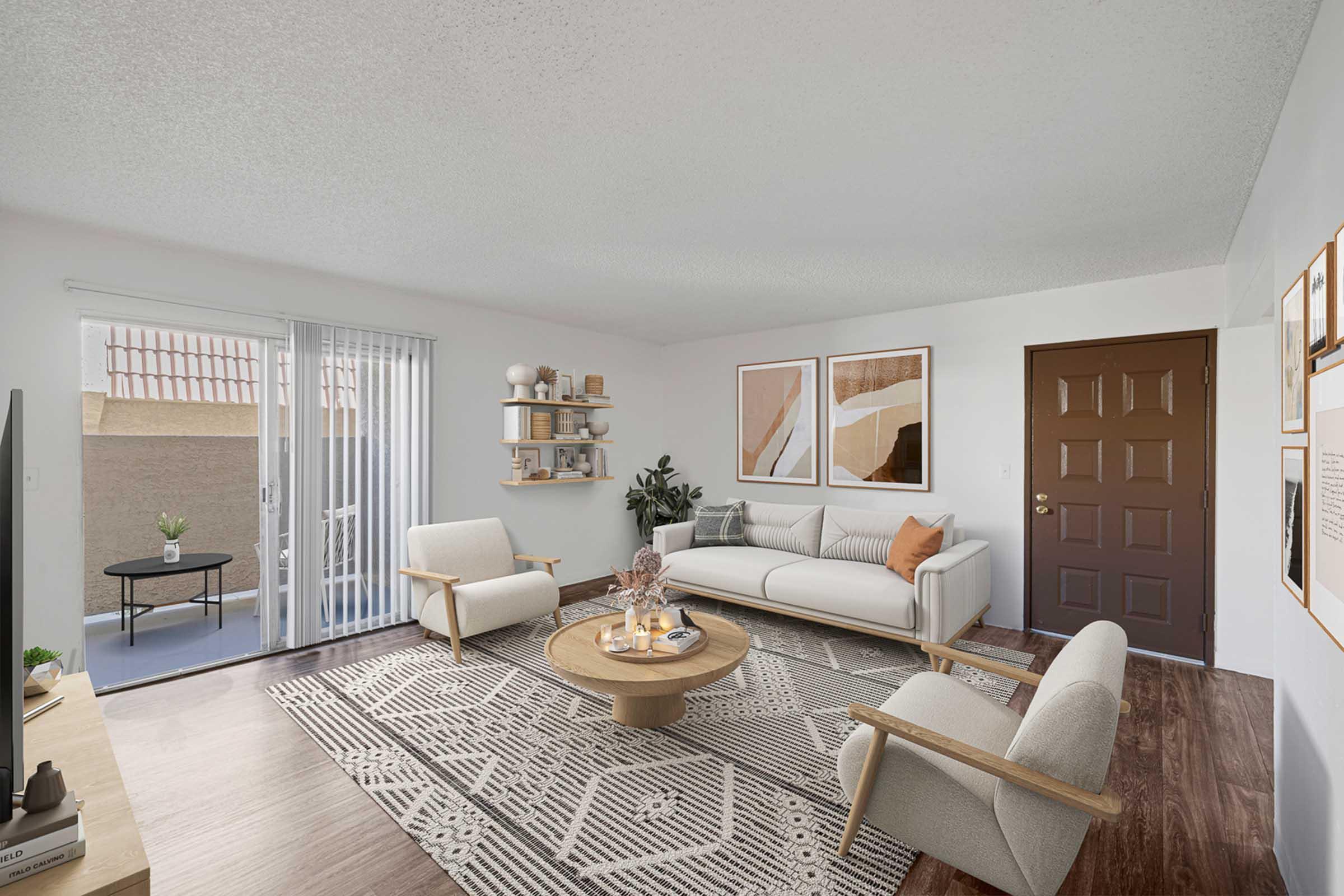 A modern living room featuring a light-colored sofa with orange and beige accents, two armchairs, a round coffee table, and a patterned area rug. There are framed artworks on the walls, a decorative plant, and a door leading outside to a patio area. Natural light fills the space.