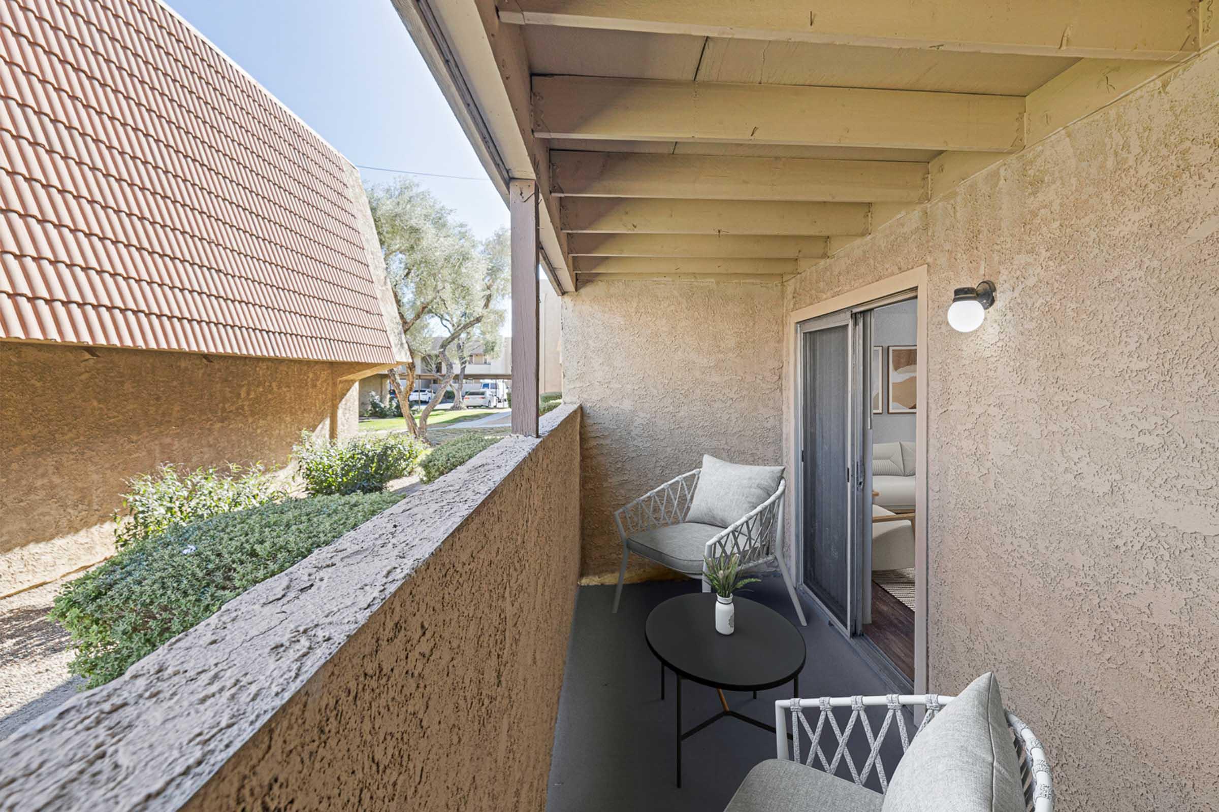 A cozy balcony with two chairs and a small table, surrounded by textured walls, offering a view of nearby greenery and residential buildings. Natural light filters in, creating a warm atmosphere.