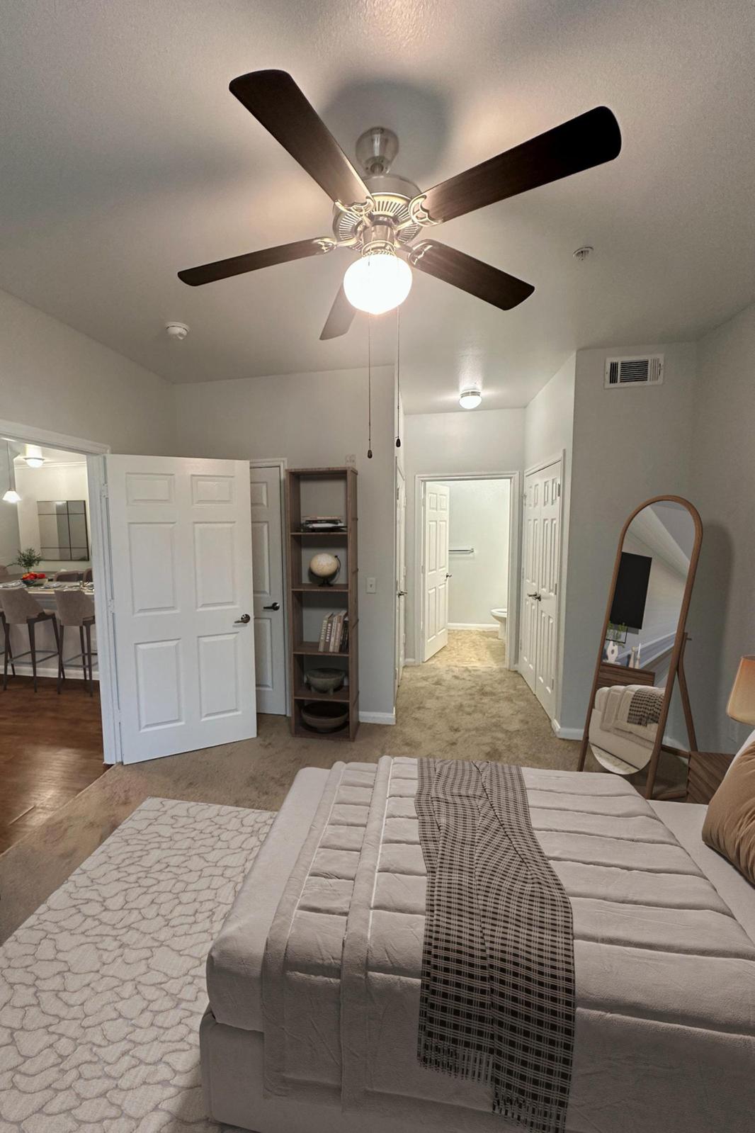 A spacious bedroom featuring a neatly made bed with a light-colored blanket, a ceiling fan, and a large mirror. The room includes a small shelf and leads to a hallway with closed doors. Soft lighting enhances the inviting atmosphere, with a cozy carpeted floor throughout.