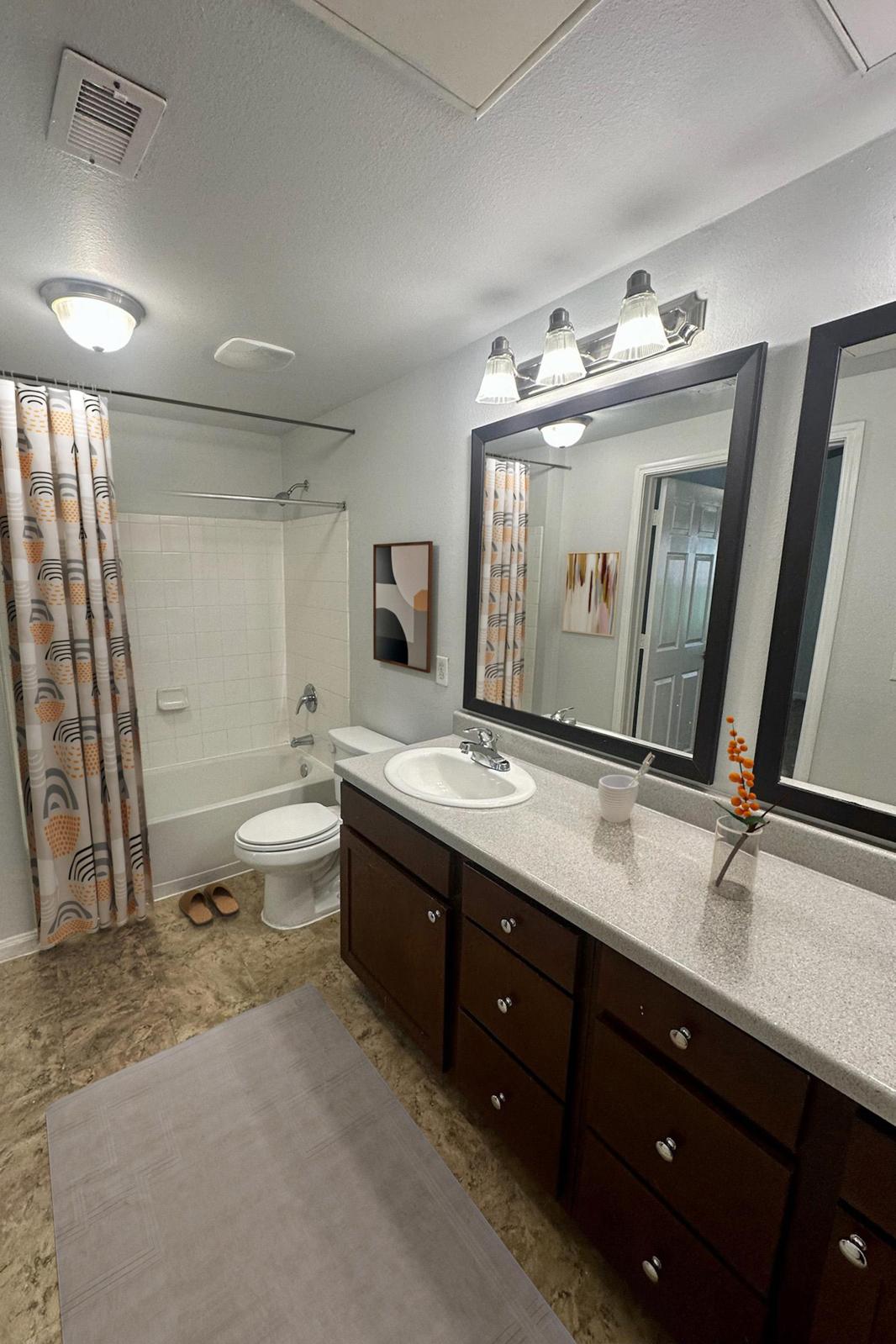 Modern bathroom with a beige and gray color scheme. Features a dual sink vanity with dark wood cabinets, a large mirror, and stylish lighting. Includes a bathtub with a shower, an orange and cream curtain, a rug on the floor, and decorative elements like a small vase with flowers.
