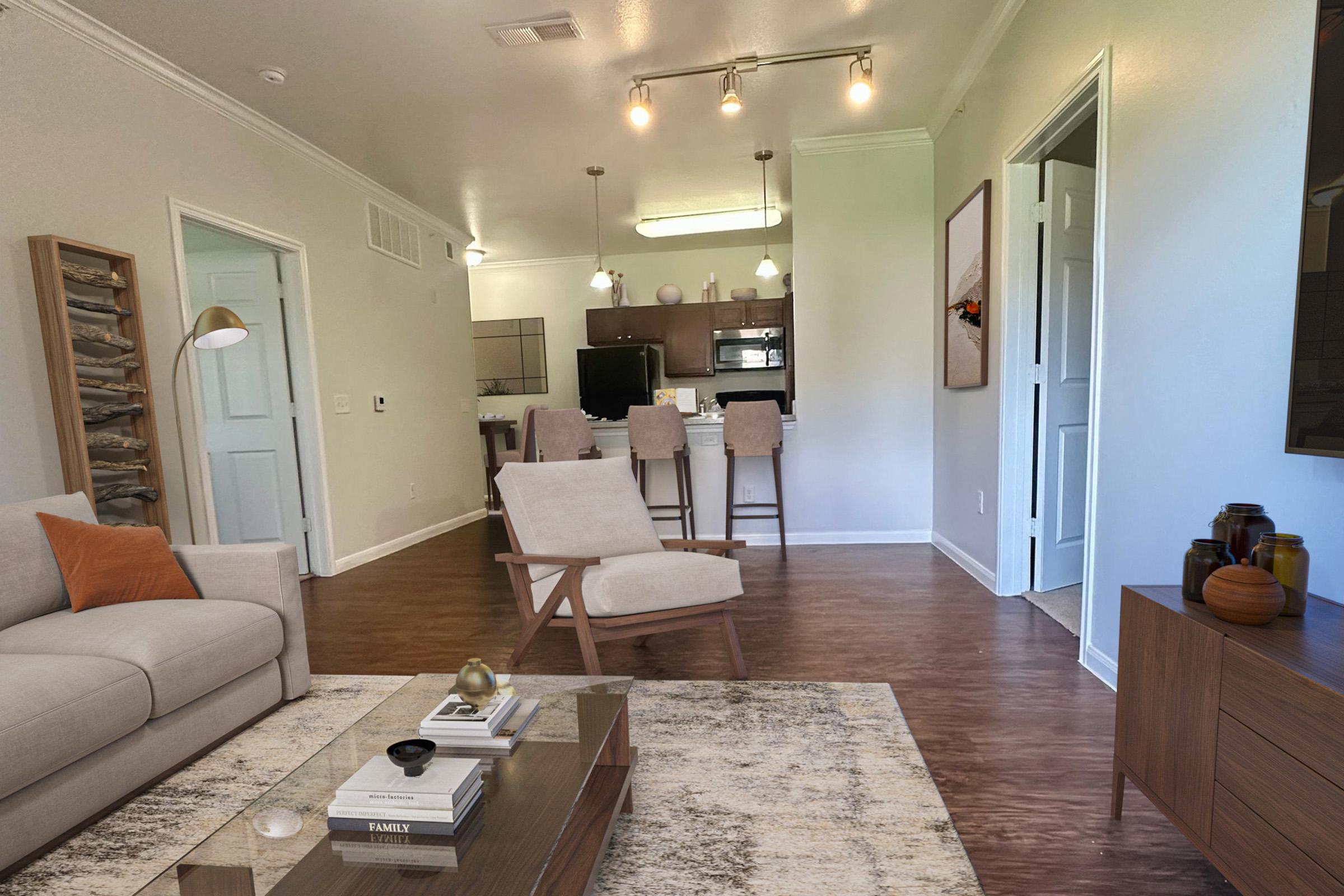 Modern living room with a light-colored sofa, a wooden coffee table on a plush rug, and a cozy armchair. There is a kitchen area in the background with bar seating, and contemporary lighting fixtures. The walls are painted in neutral tones, creating a bright and inviting atmosphere.