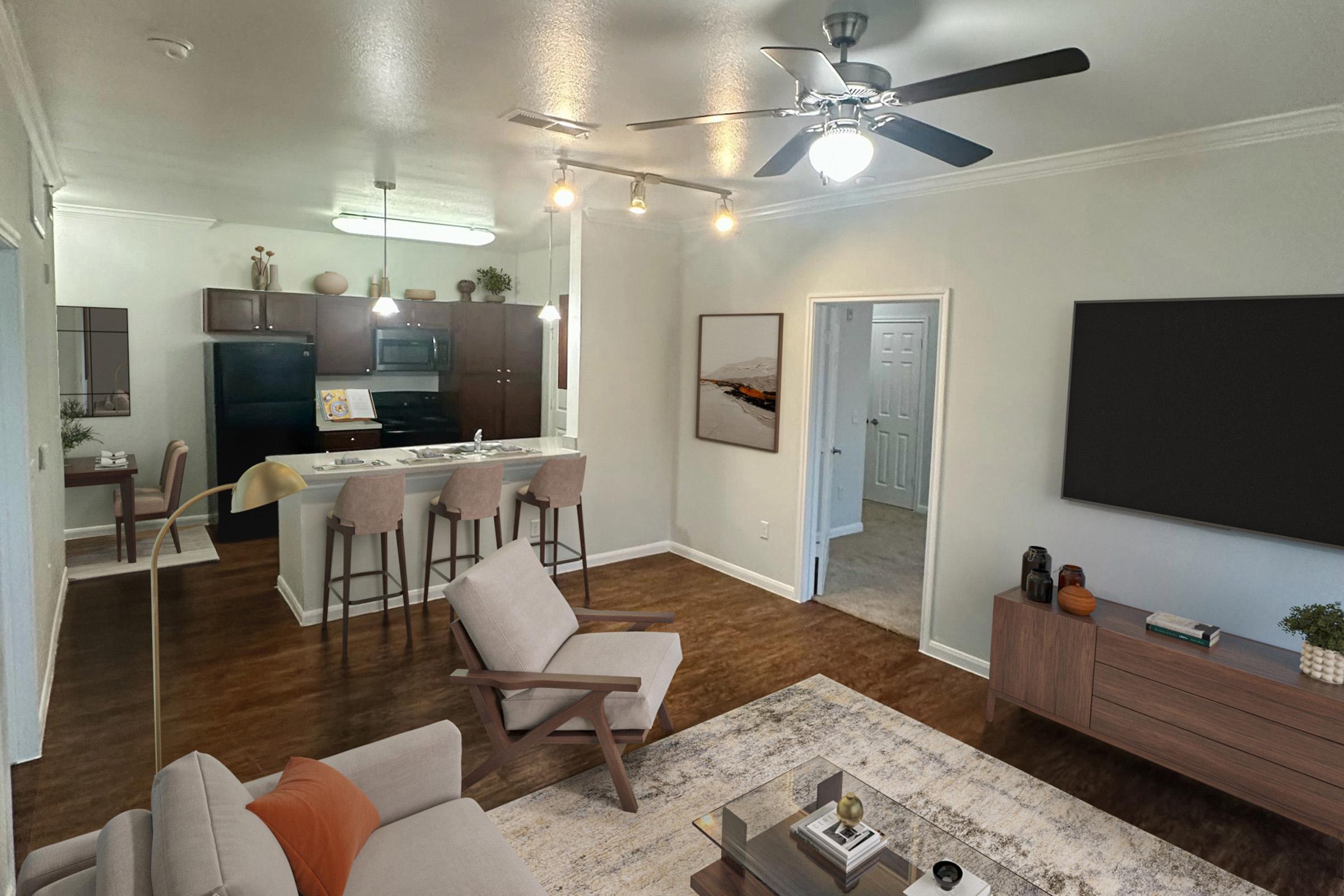 A modern living space featuring a comfortable light-colored armchair and sofa, a large area rug, and a wooden coffee table. In the background, there's an open kitchen with dark cabinets, a black refrigerator, and bar stools. A ceiling fan and wall-mounted TV enhance the ambiance. Light streams through a doorway.