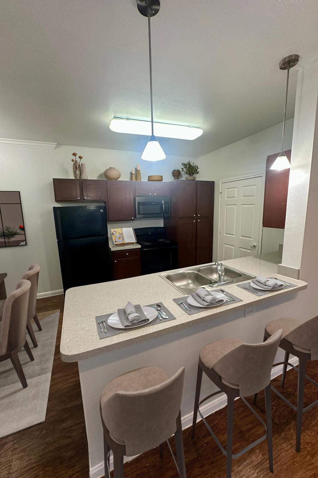 Modern kitchen with dark wood cabinets, a black refrigerator, and a stove. There are two pendant lights above a countertop with two place settings. In the background, a dining area features a round table with gray upholstered chairs. The floor is dark wood, and the walls are painted white, creating a bright atmosphere.