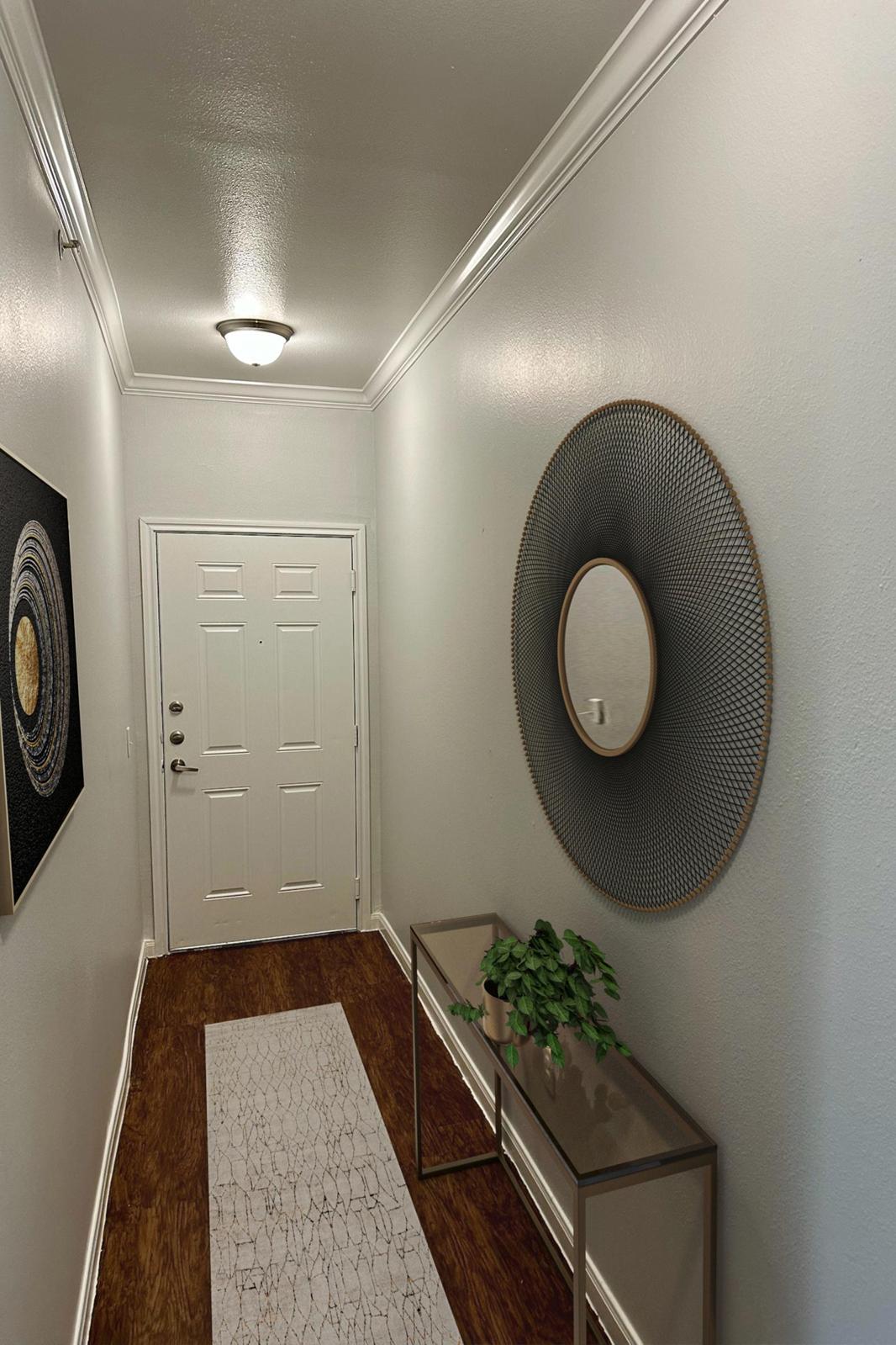 A well-lit hallway featuring a white wall and light wood flooring. On the left, a round mirror with a decorative frame hangs next to an abstract wall art piece. A console table with a potted plant is positioned on the right, complemented by a long, narrow runner rug along the hallway's length. The front door is visible at the end.