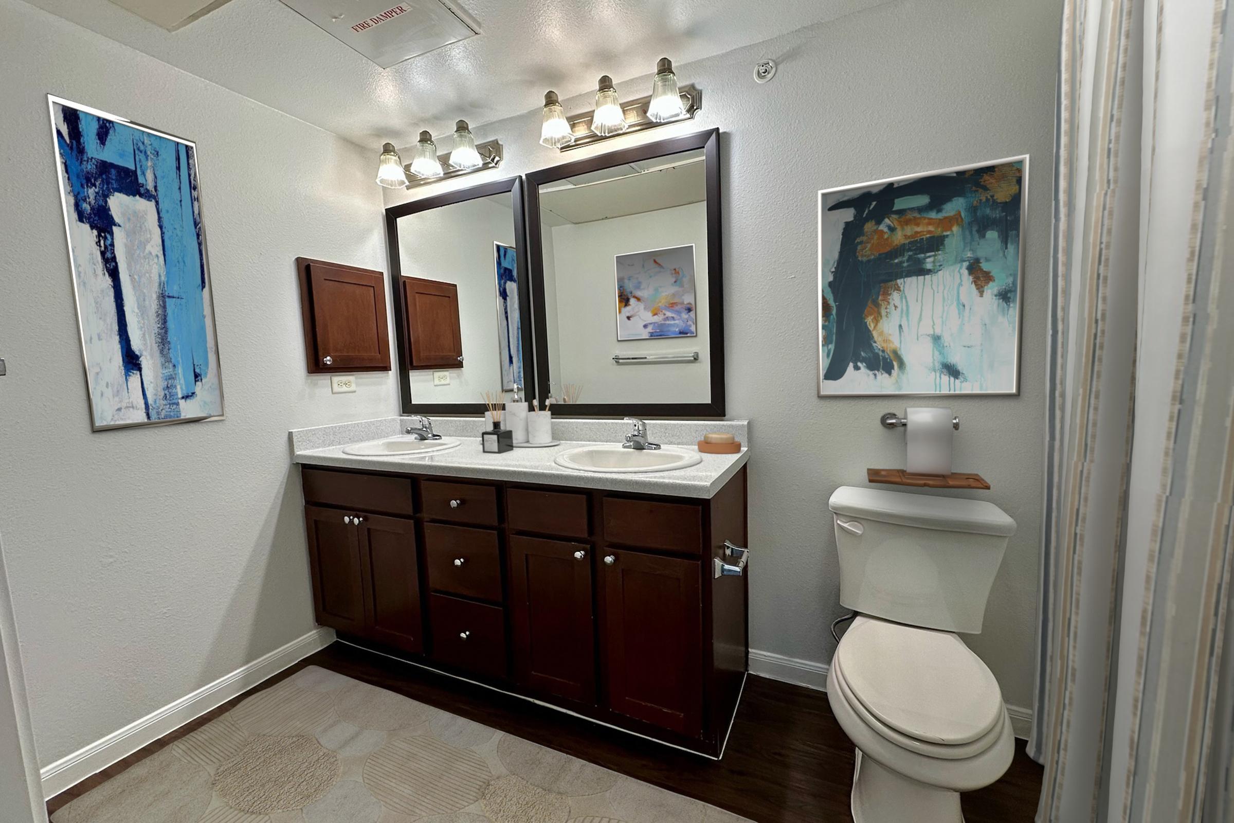 A well-lit bathroom featuring a double vanity with dark wood cabinets, a large mirror above, and contemporary artwork on the walls. There is a white toilet to the right, soft flooring, and towel hooks. The overall color scheme is neutral and inviting.