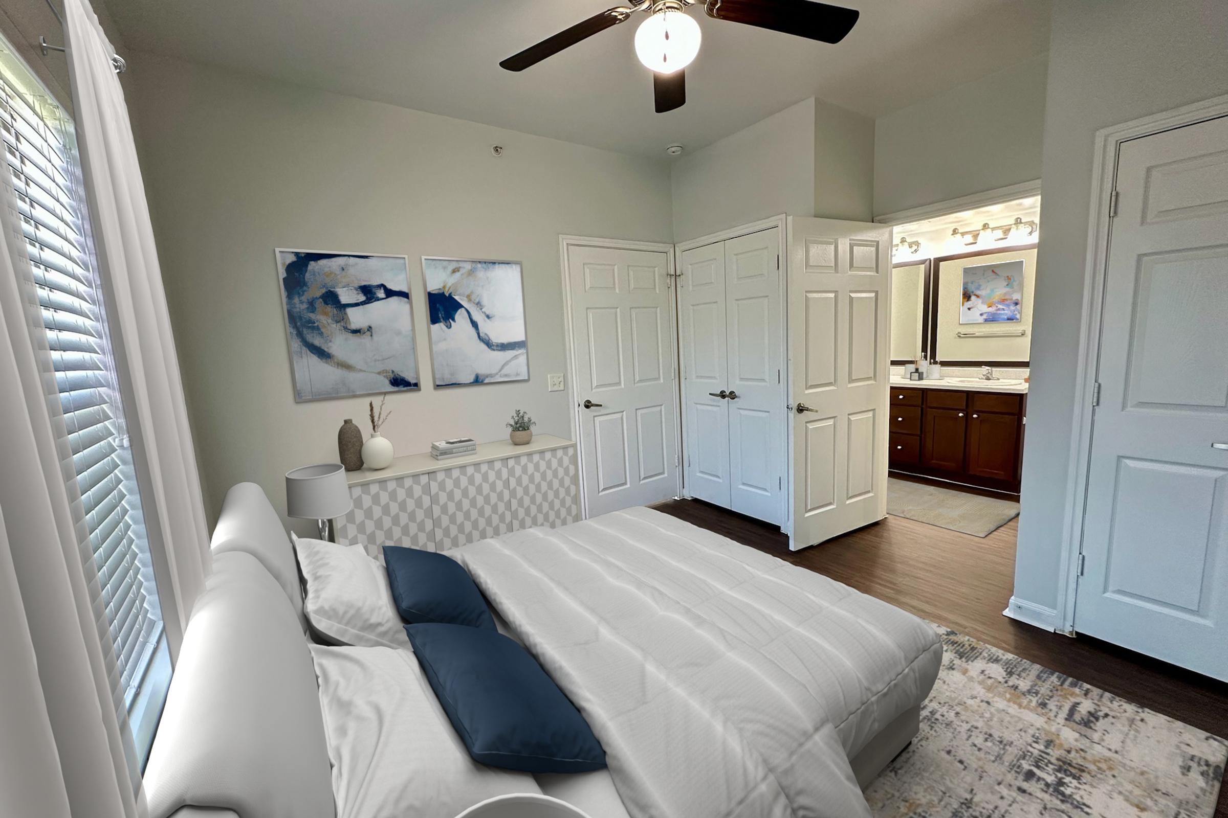 A well-lit bedroom with a modern design featuring a white bed with blue pillows, a textured gray accent wall, and abstract artwork. A nightstand and decorative items are placed on a dresser. The room includes light-colored walls and hardwood flooring, with a doorway leading to a bathroom.