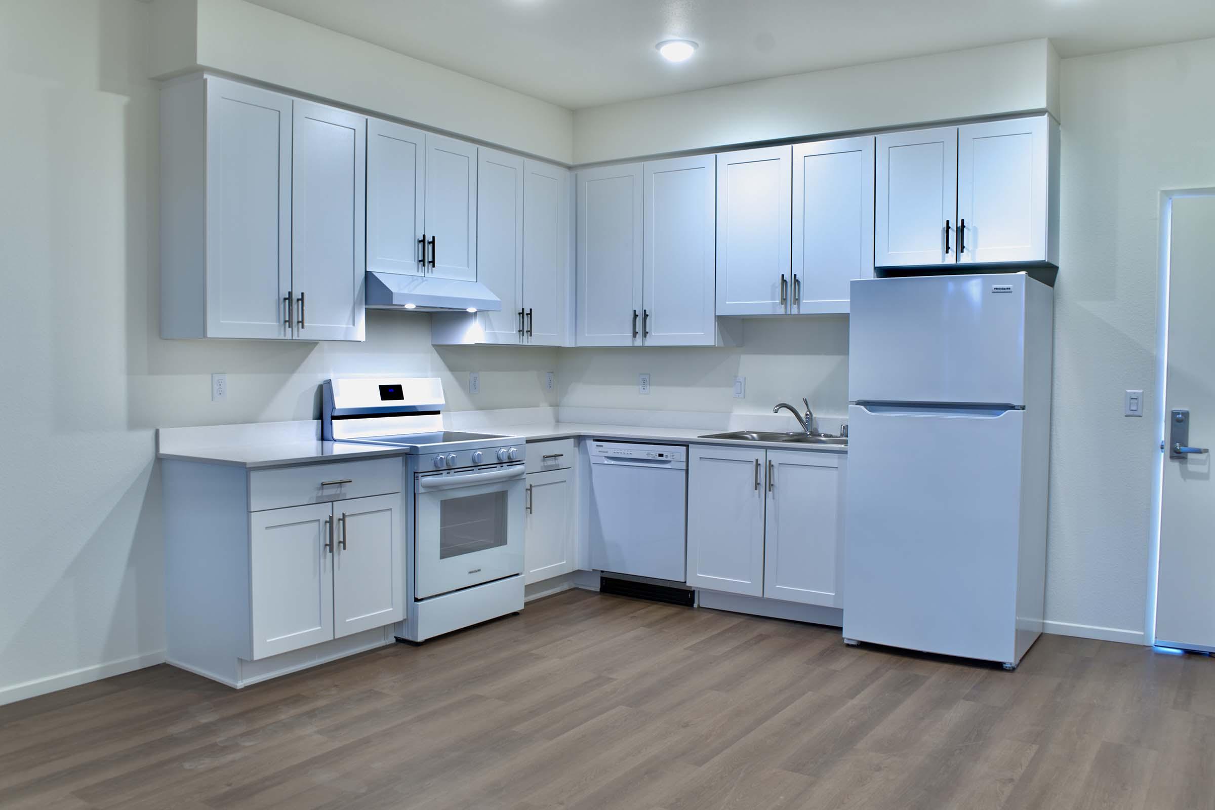 Modern kitchen with white cabinetry, stainless steel appliances including a stove, oven, refrigerator, and dishwasher, set against light-colored walls and wooden flooring. The space is bright and airy, featuring under-cabinet lighting and a clean, minimalist design.
