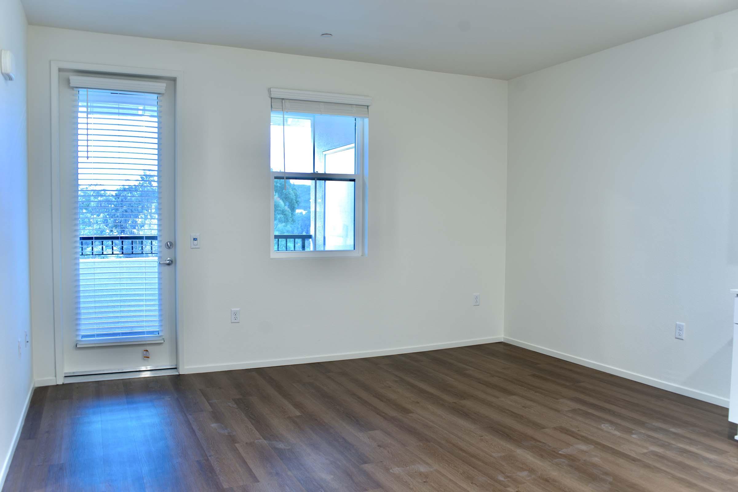 Empty room with light-colored walls and wooden flooring. A sliding glass door leads to a small balcony, with a window adjacent to it allowing natural light to enter. The space is unfurnished, creating a bright and open atmosphere.