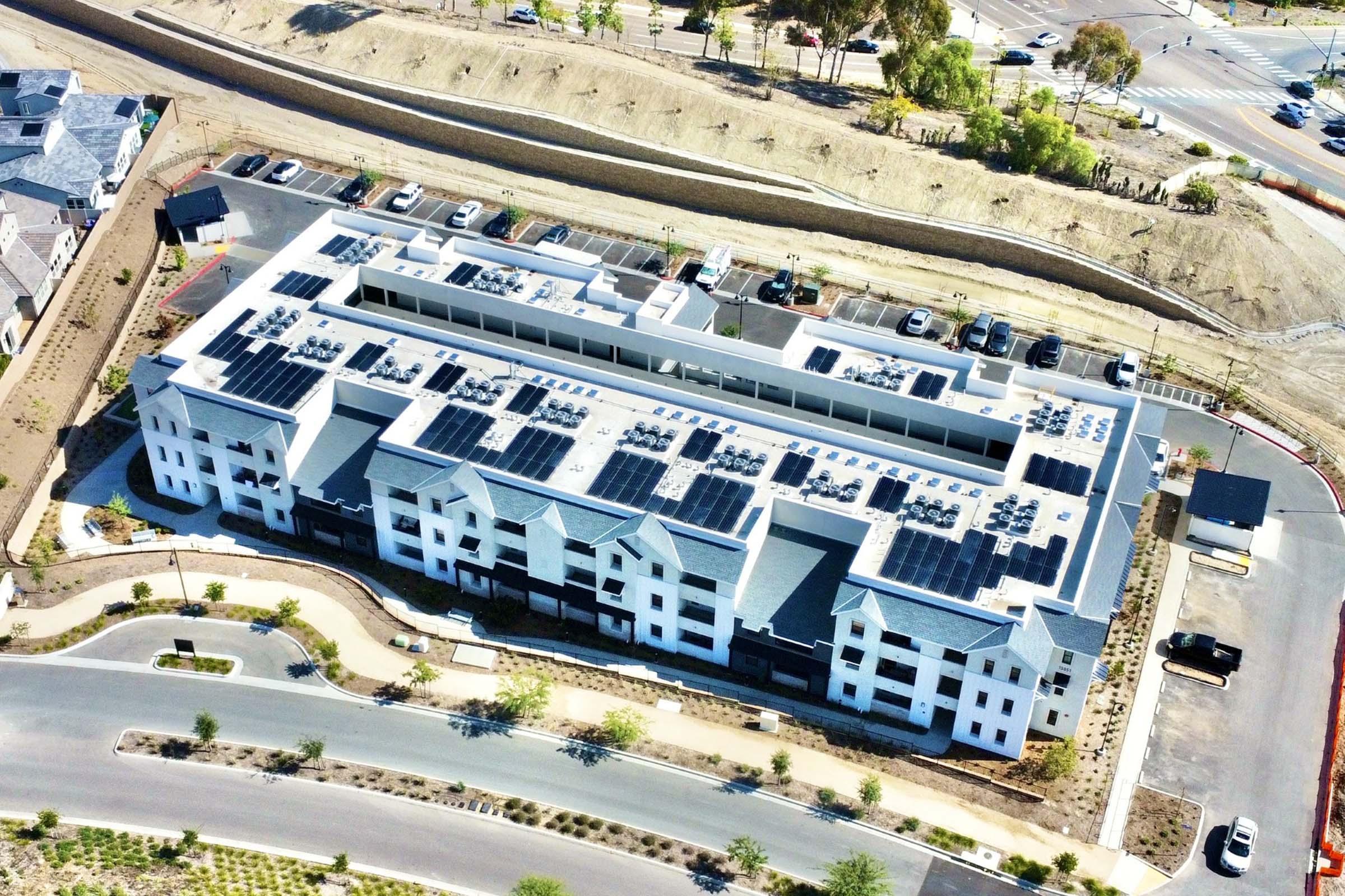 Aerial view of a modern multi-story residential building featuring a flat roof covered with solar panels. Surrounding the building are landscaped areas and parking spaces. Nearby, there is a road and undeveloped land, indicating an expanding urban area.