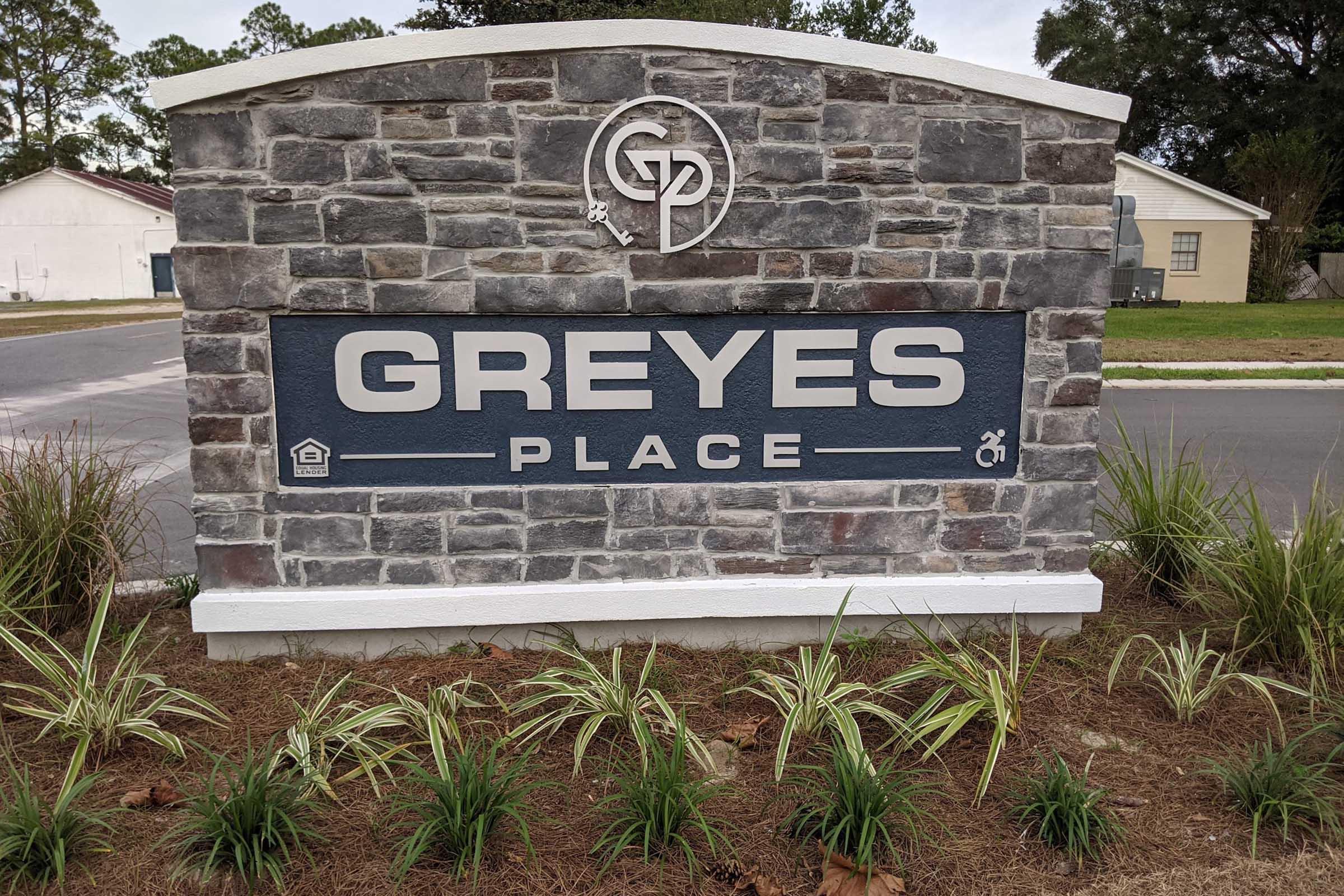Sign for "Greyes Place" featuring a stone design, with the letters prominently displayed in white against a dark blue background. The sign includes a logo with the initials "GP" and symbols for amenities, surrounded by landscaped greenery.