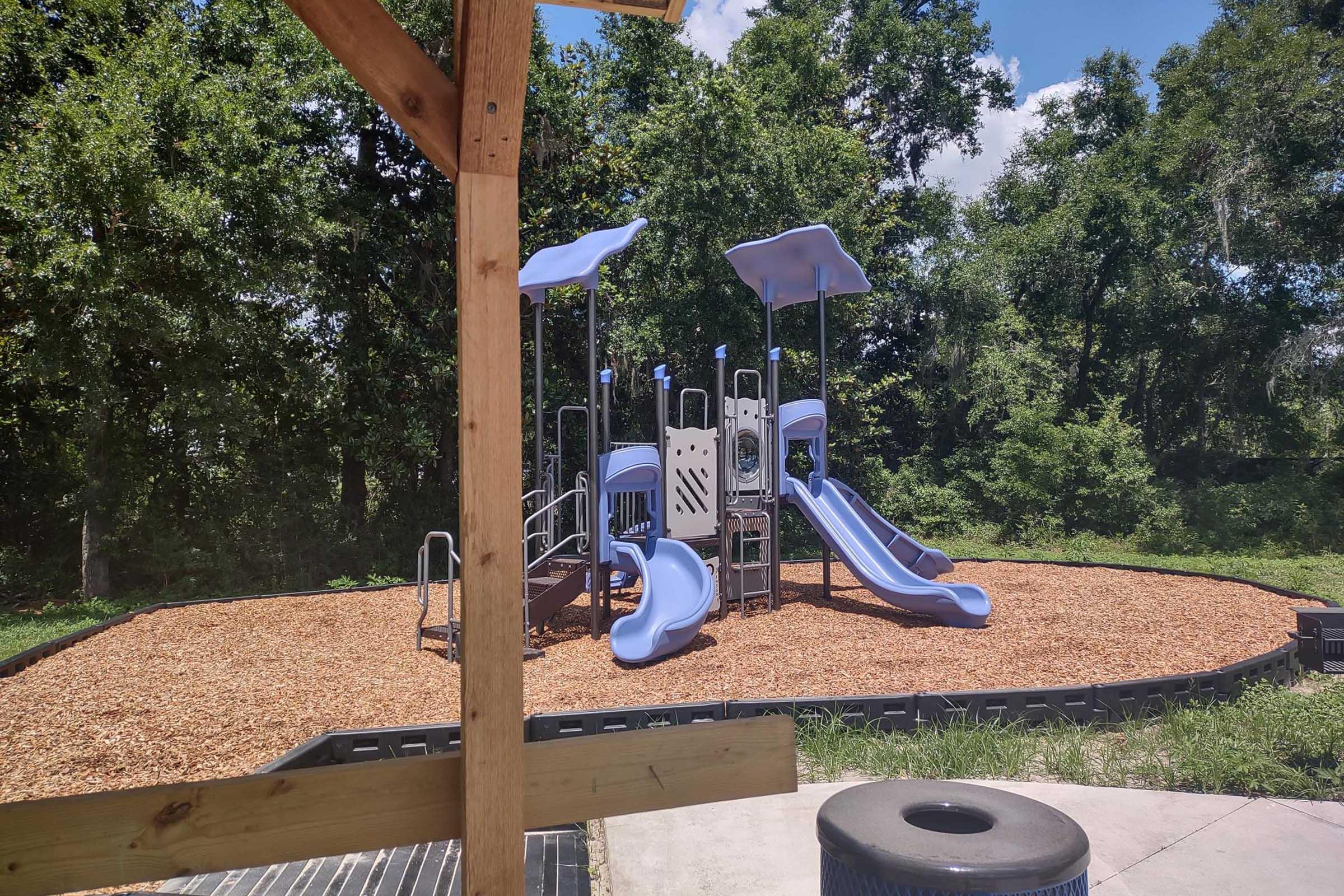 Playground area featuring a modern play structure with two blue slides. The ground is covered with wood chips, and the space is surrounded by trees under a clear blue sky. A wooden frame partially obscures the view, suggesting it is taken from a nearby shelter or structure.