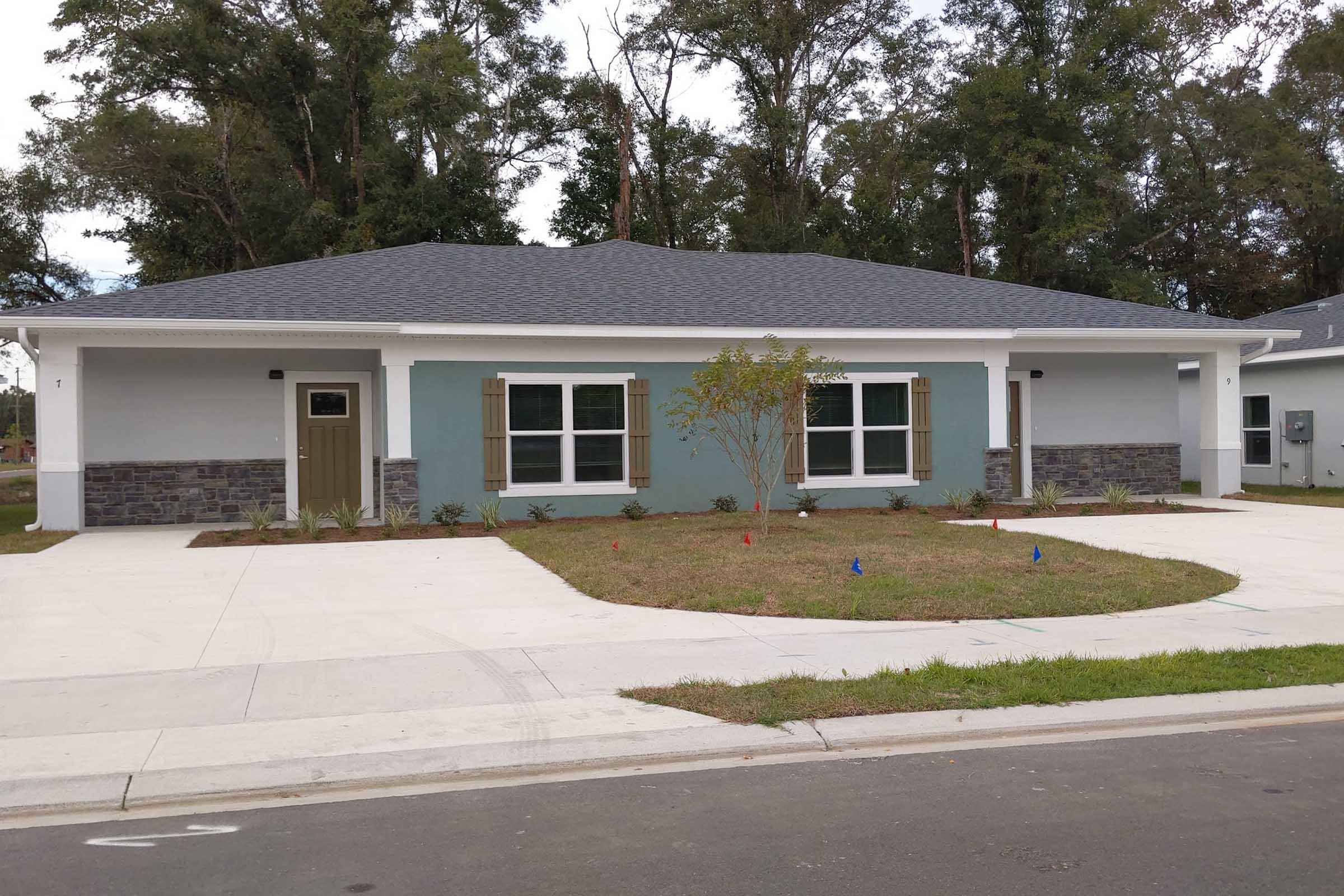 A modern single-story duplex with a light blue exterior, stone accents, and decorative shutters. The property features a small front yard with grass and a driveway. Surrounding trees provide a natural backdrop, and there are colorful markers indicating landscaping or construction.