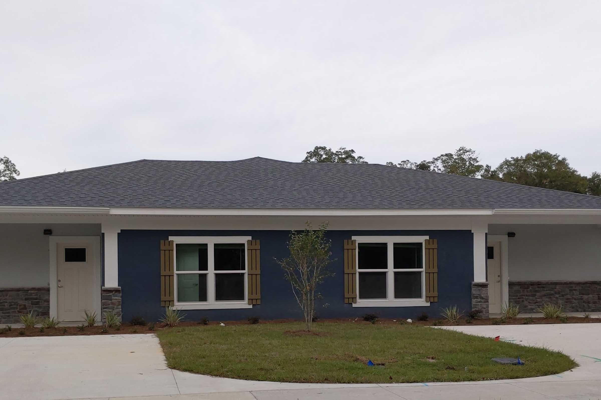 A modern one-story building with a blue exterior, white trim, and dark shutters. The front features two doors, large windows with square panes, and a small tree planted in front. The area has freshly landscaped grass and is surrounded by a paved path.