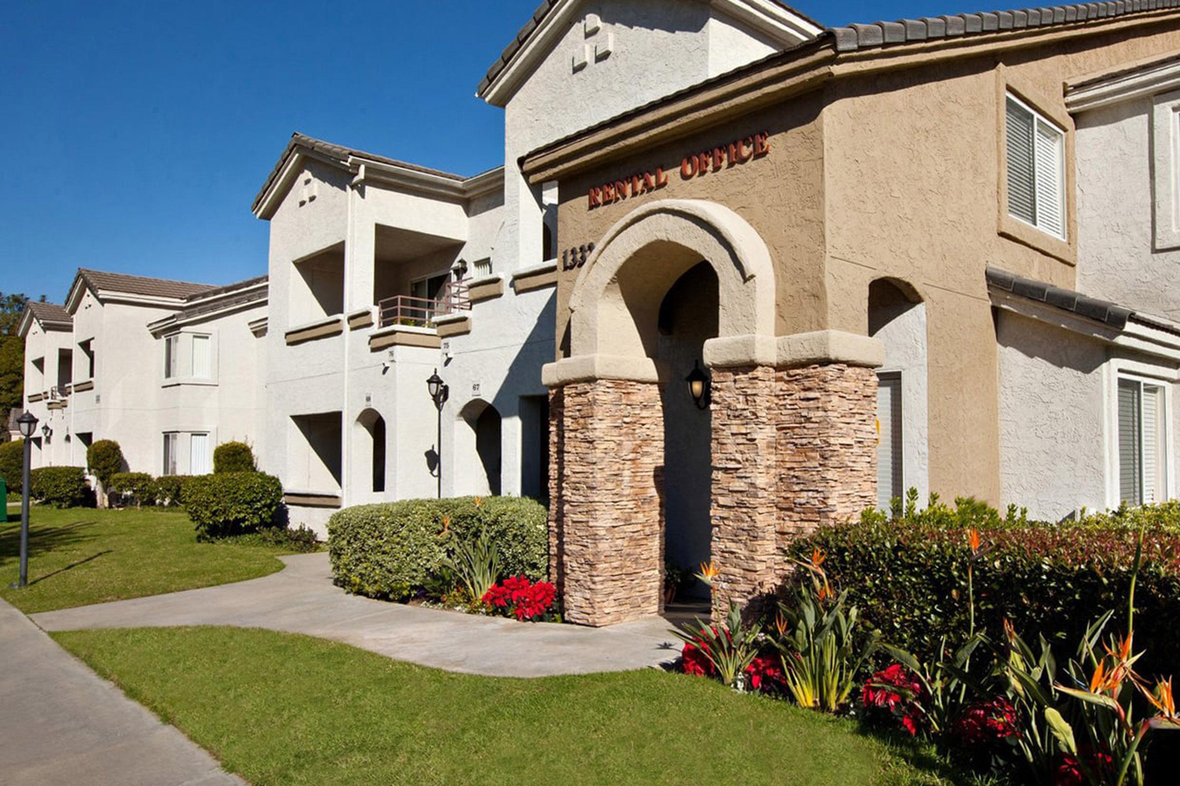 Exterior view of a residential building featuring a prominently labeled dental office. The structure has a light-colored facade, decorative stone accents, and well-maintained landscaping with colorful flowers and shrubs. Clear blue sky above enhances the inviting atmosphere.