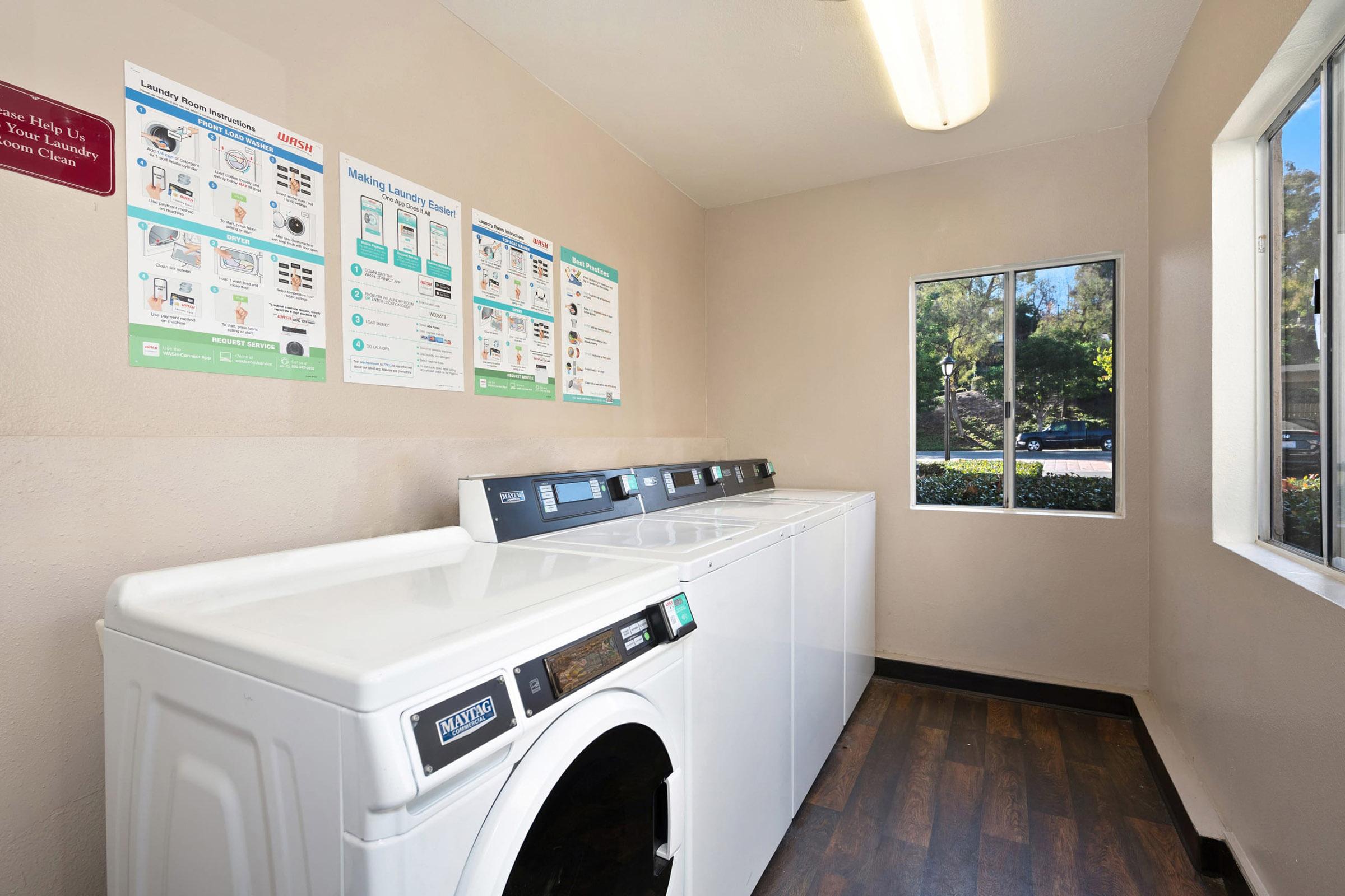 A laundry room featuring two white washing machines and a dryer, with a window showing greenery outside. Informational posters on the wall provide instructions for using the machines. The floor has a dark wood-like finish, and overhead lighting is bright.