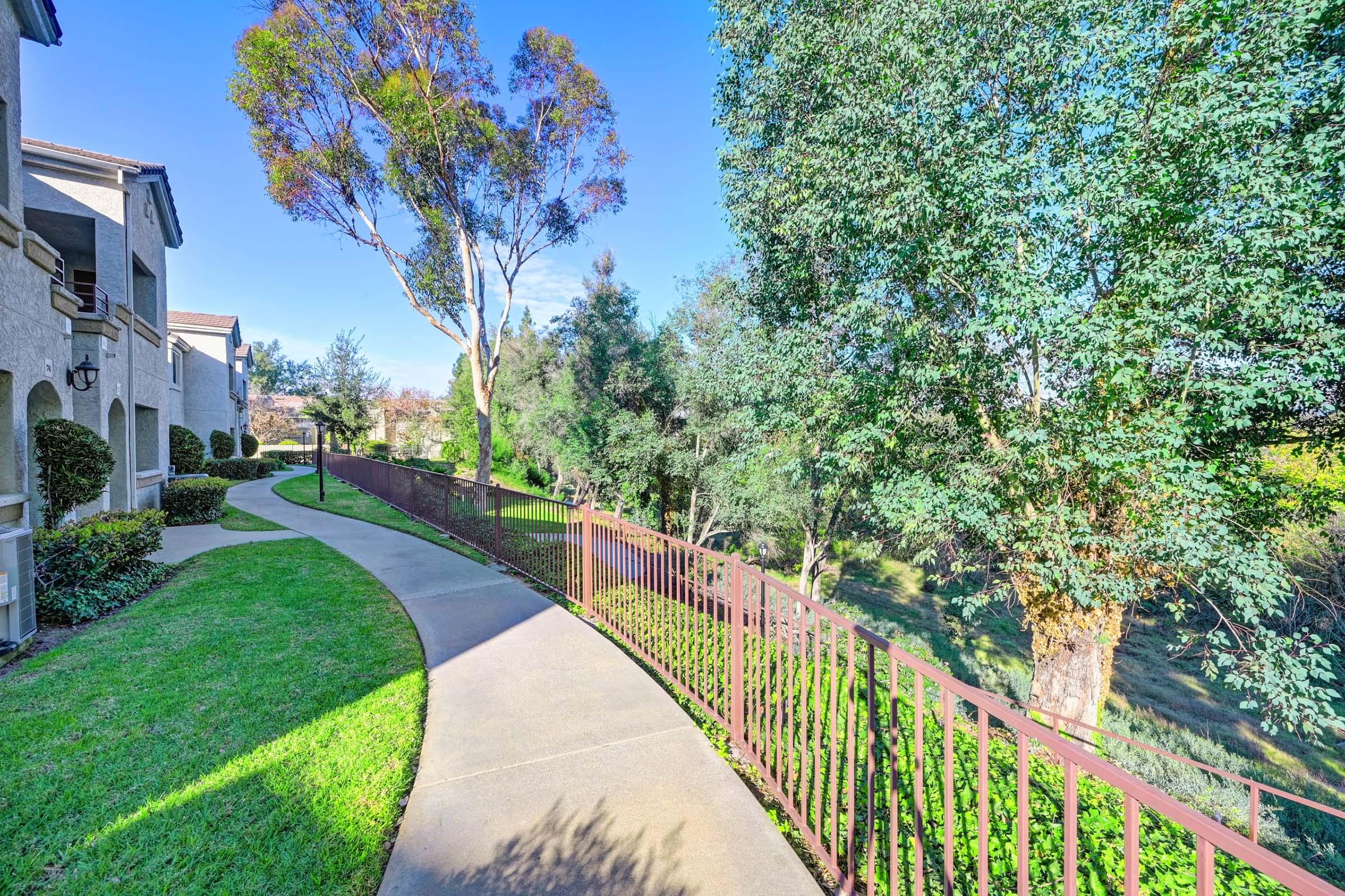 A paved walkway lined with green grass and bushes, leading alongside a brown metal fence. Lush trees and shrubs are present on both sides of the path, suggesting a peaceful, landscaped community area with clear blue skies above.