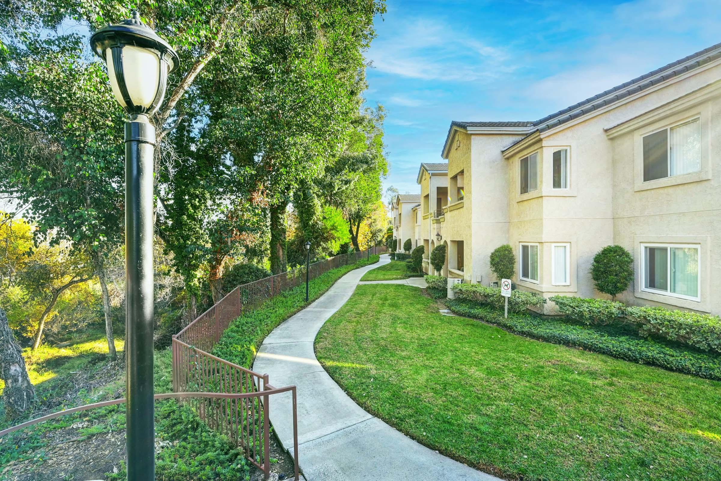 A landscaped pathway meanders through a green area beside a residential building. The path features a metal railing and is lined with trees and shrubs. A lamppost stands along the walkway, adding to the serene, suburban ambiance.
