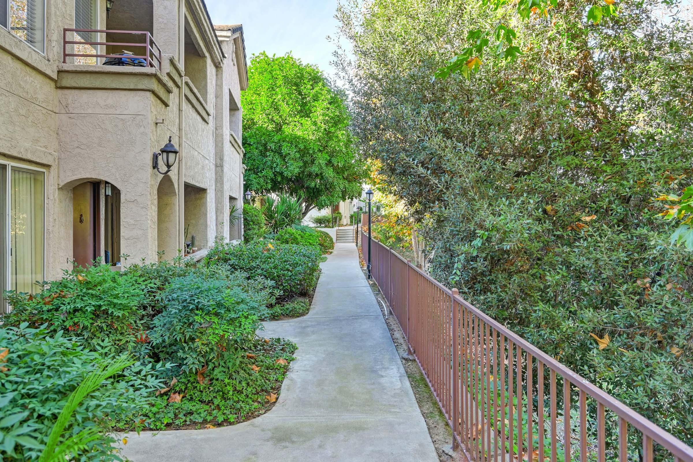 A pathway bordered by lush greenery leads along a beige building with arched doorways and windows. A wrought-iron fence runs adjacent to the walkway, separating it from a dense row of shrubs and trees. Sunlight filters through, illuminating the serene outdoor space.
