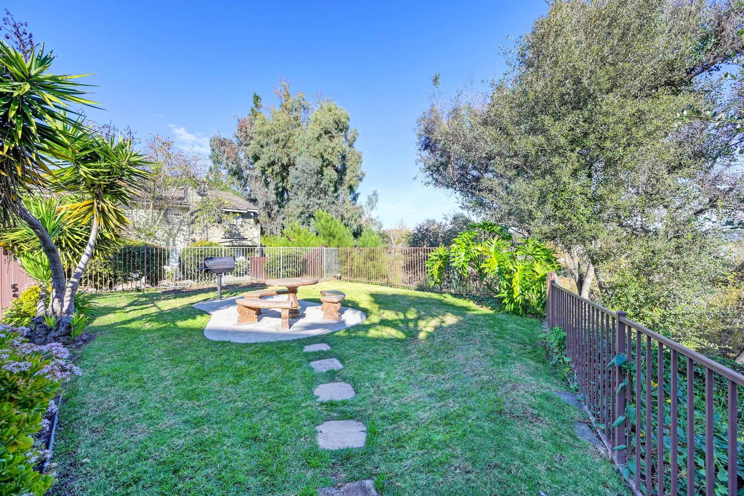 A serene backyard scene featuring a well-maintained lawn, stone pathway, and a circular seating area with chairs around a fire pit. Lush trees and greenery surround the space, offering a peaceful atmosphere, with a clear blue sky overhead.