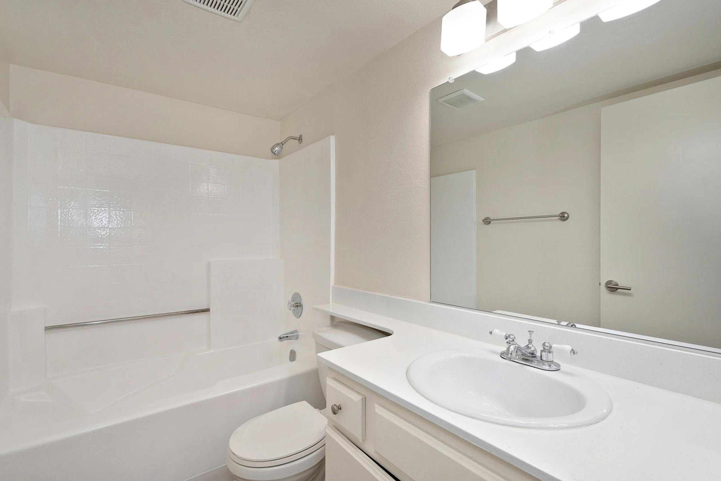 A clean and simple bathroom featuring a white bathtub with a shower, a sink with a mirror above it, and neutral-colored walls. The space includes a towel bar and modern lighting fixtures, creating a bright and inviting atmosphere.