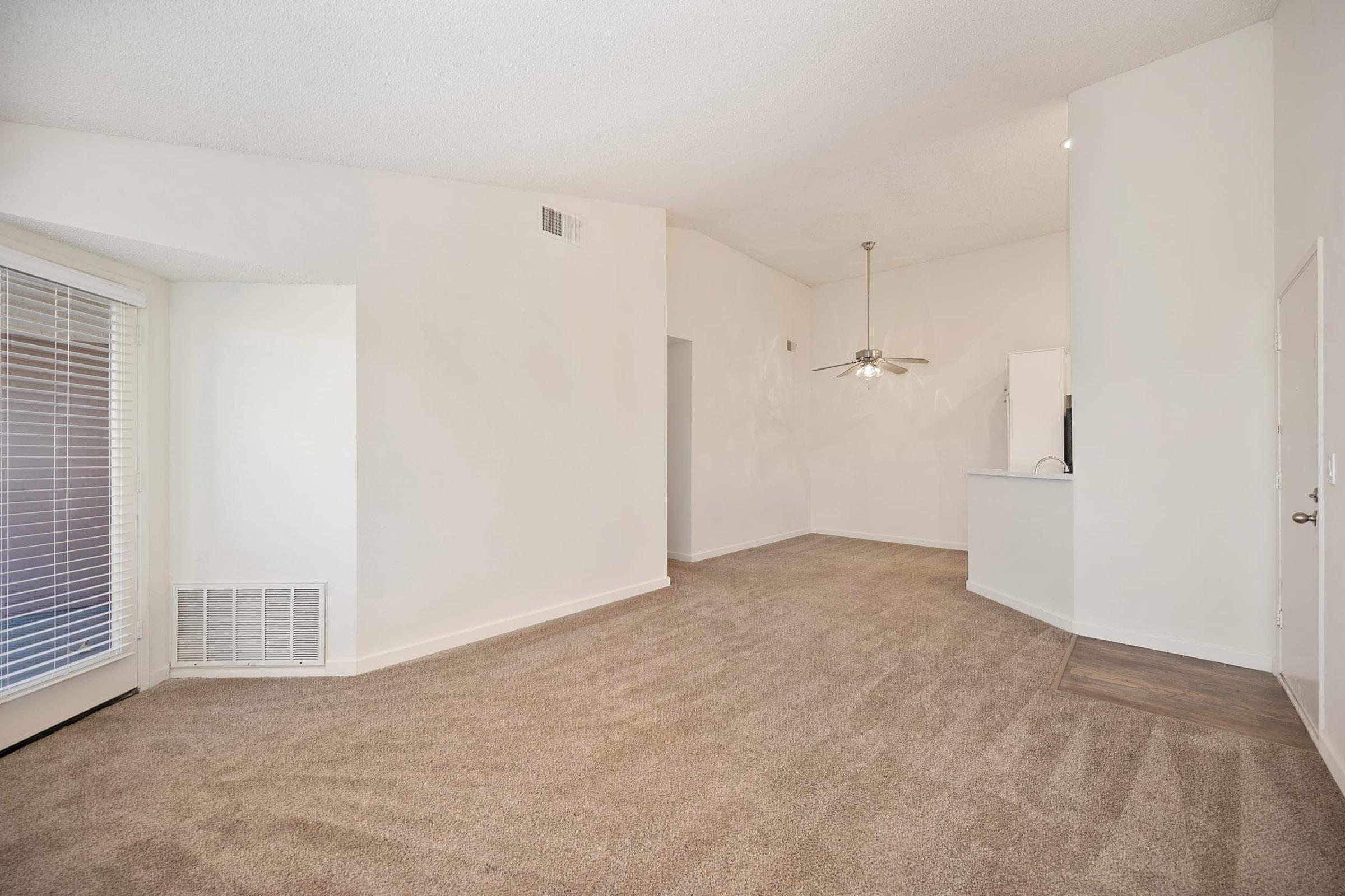Spacious, empty living room with light beige carpet and white walls. Large window with blinds provides natural light, and a ceiling fan is visible. The room leads to a kitchen area, creating an open-concept feel.