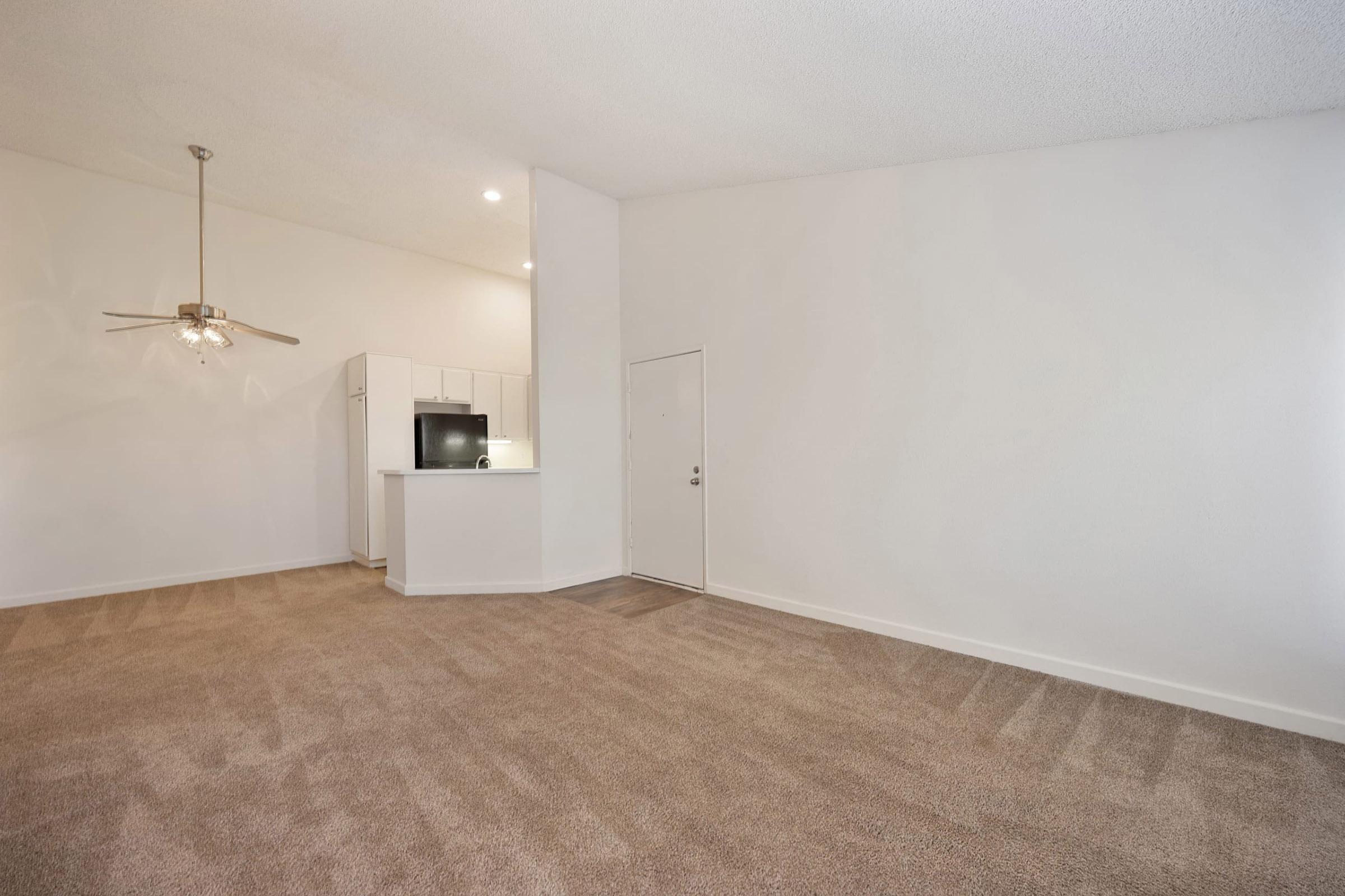 A spacious, empty living area with beige carpet and white walls. To the left, a kitchen area is visible, featuring a refrigerator and a small countertop. A ceiling fan is mounted above, and there's a main door at the front of the room, leading outside. The natural light enters from large windows, creating a bright atmosphere.