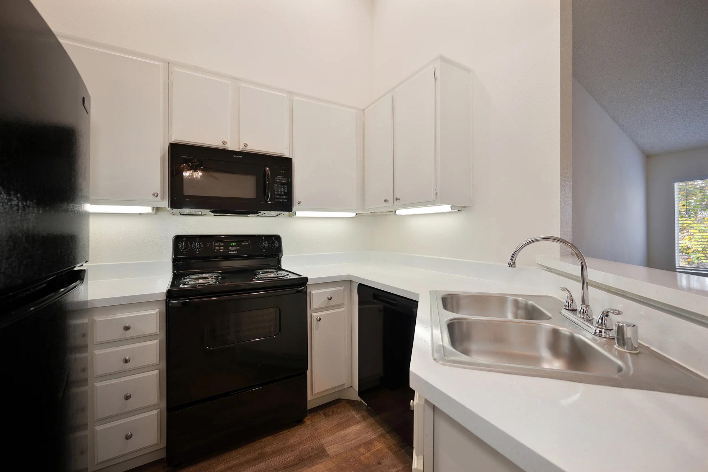 Modern kitchen featuring black appliances, including a refrigerator, microwave, and stove. The cabinetry is white with a sleek countertop, and there are dual sinks. Soft lighting illuminates the space, showcasing a clean and organized layout with wooden flooring.