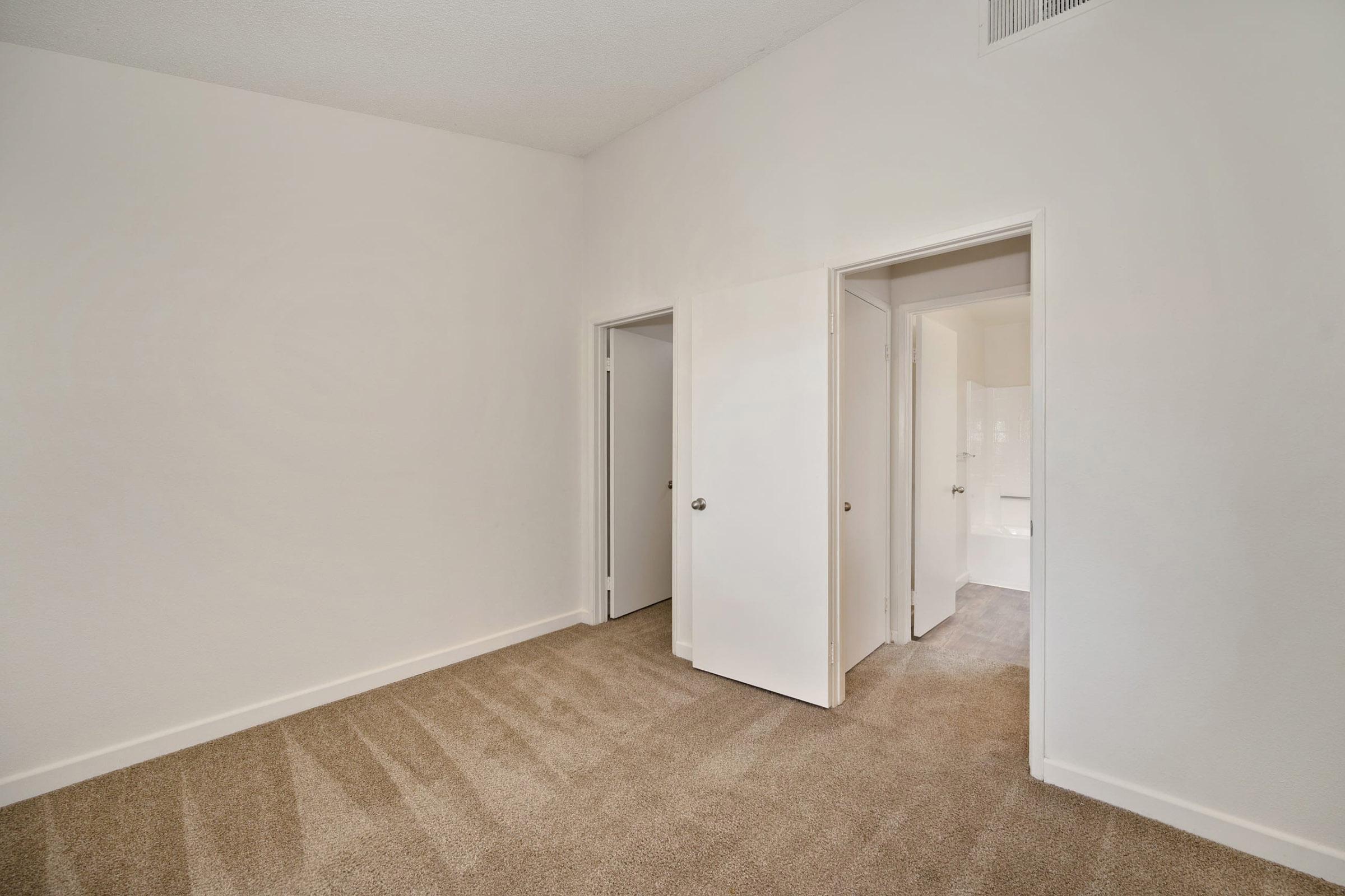 A bright, empty room featuring light-colored walls and plush beige carpet. There are two closed doors leading to adjoining spaces, and ample natural light enters the room. The ceiling is high, contributing to an airy feeling, and there are no furnishings or decor present.