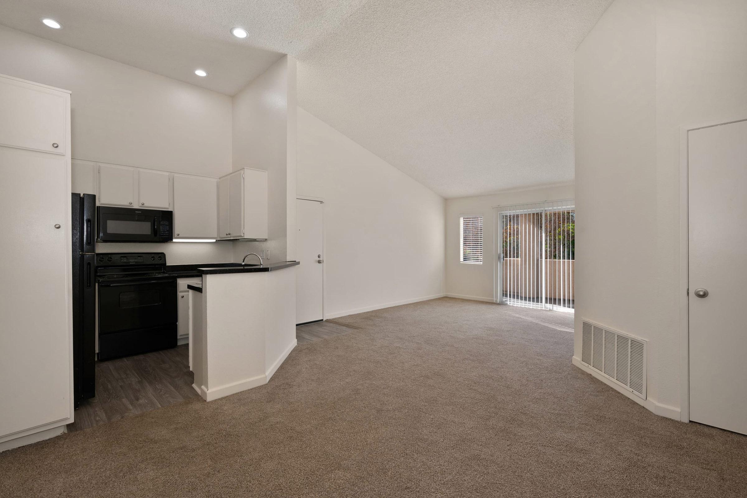Interior view of a modern, spacious living area featuring light beige carpet and white walls. On the left, a kitchen with black appliances and cabinetry. Large windows allow natural light, and a sliding door leads to an outside area. The high ceiling adds to the room's openness.