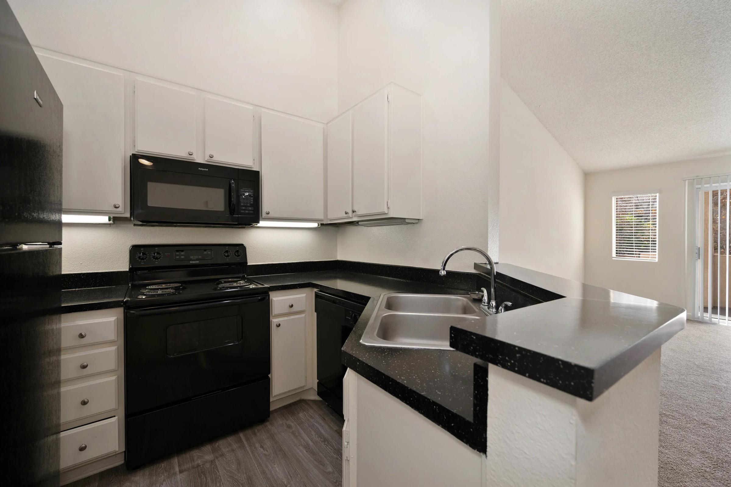 A modern kitchen featuring white cabinets, black appliances including a stove, microwave, and refrigerator. The countertop is dark and includes a sink, with a view of a carpeted area and sliding glass doors leading outside. Natural light enters through the doors, creating a bright atmosphere.